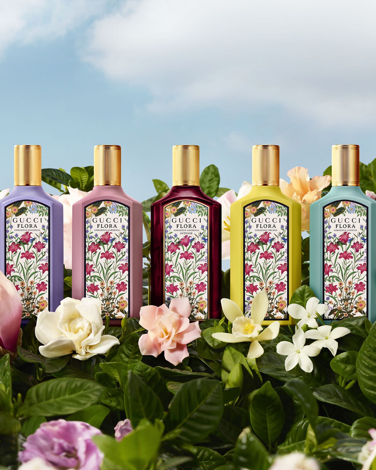 Five Gucci Flora Gorgeous bottles in lilac, pink, burgundy, yellow and turquoise, each with a floral print label, among gardenias and white blossoms under a blue sky