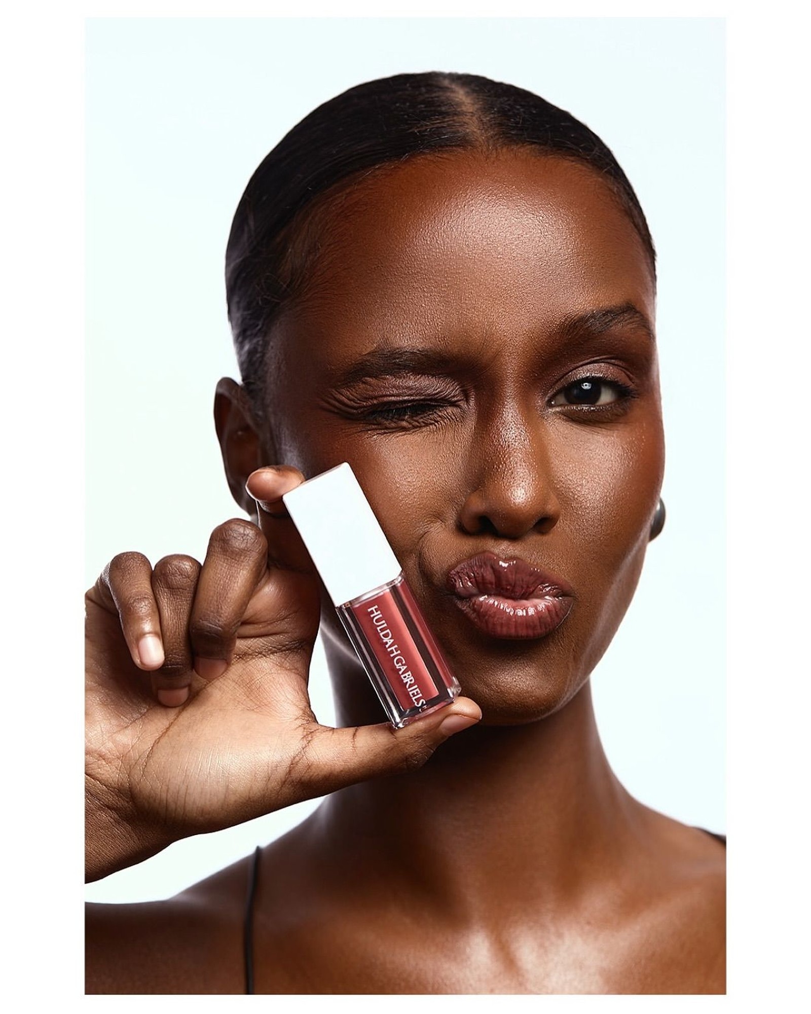 Model with a center part and a playful wink and pout, holding a red lip gloss tube against her cheek on a white background