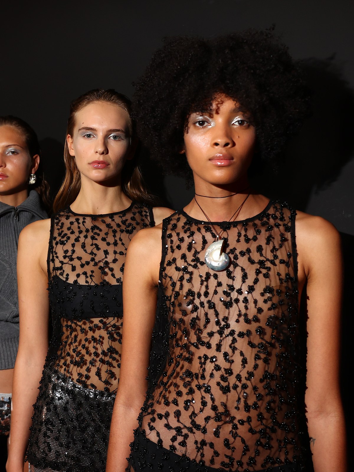 Two models backstage in sheer embellished looks and a draped knit top