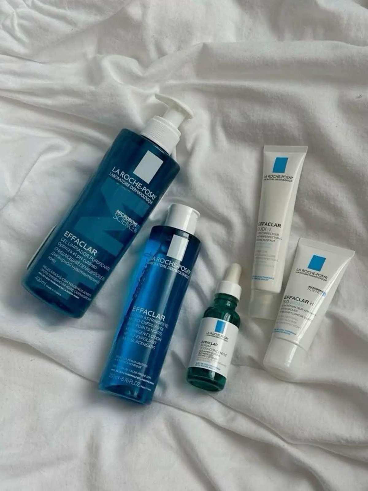 Flat lay of La Roche-Posay Effaclar cleansing gel, astringent lotion, serum and creams on white rumpled bedding