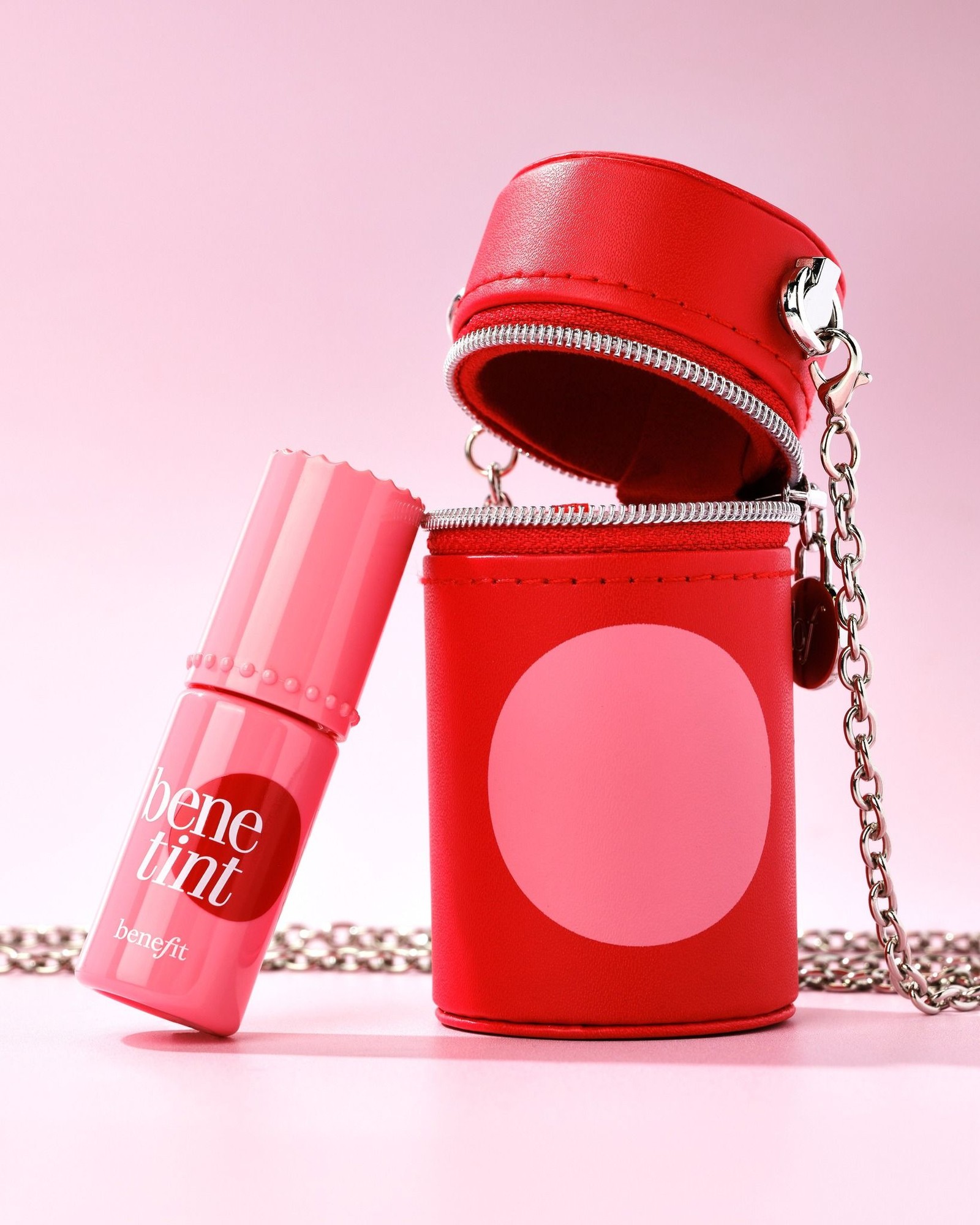 Pink Benetint bottle leaning against a red zip-up charm case with a silver chain, on a pale pink background