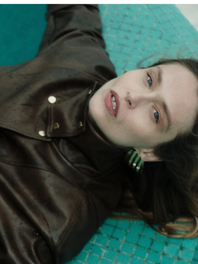 Model in a brown leather-look jacket reclining beside turquoise water