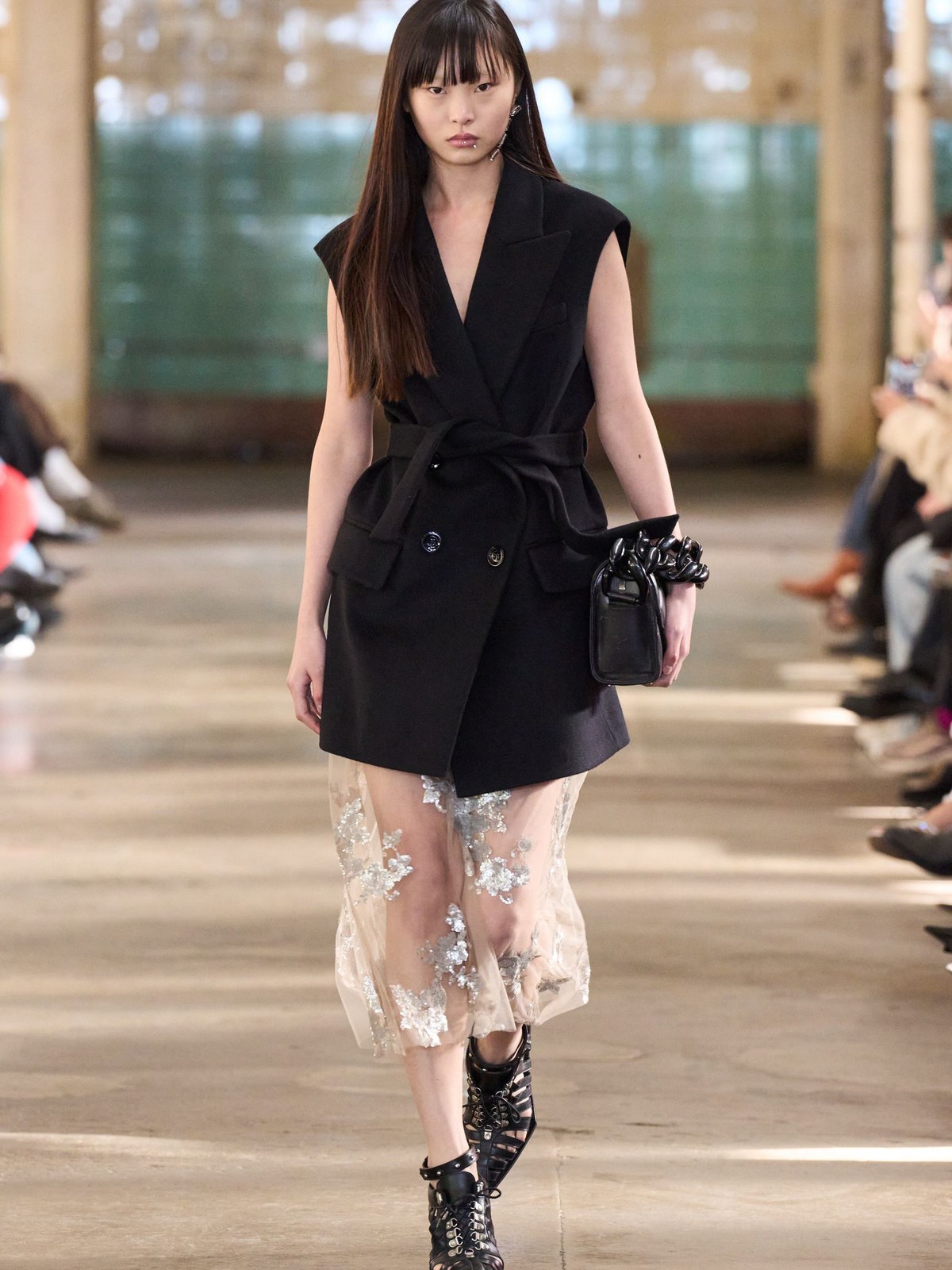 Model in a black vest and sheer embroidered trousers on the runway