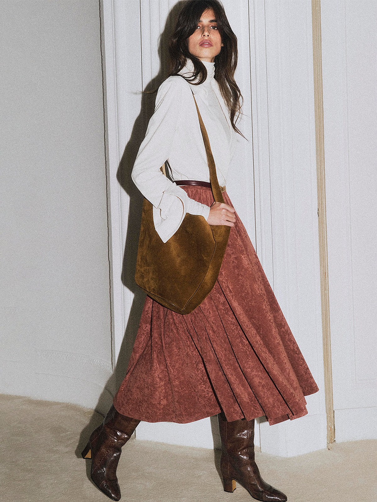 Model in a white blouse, rust pleated midi skirt and boots with a brown bag by a tall window