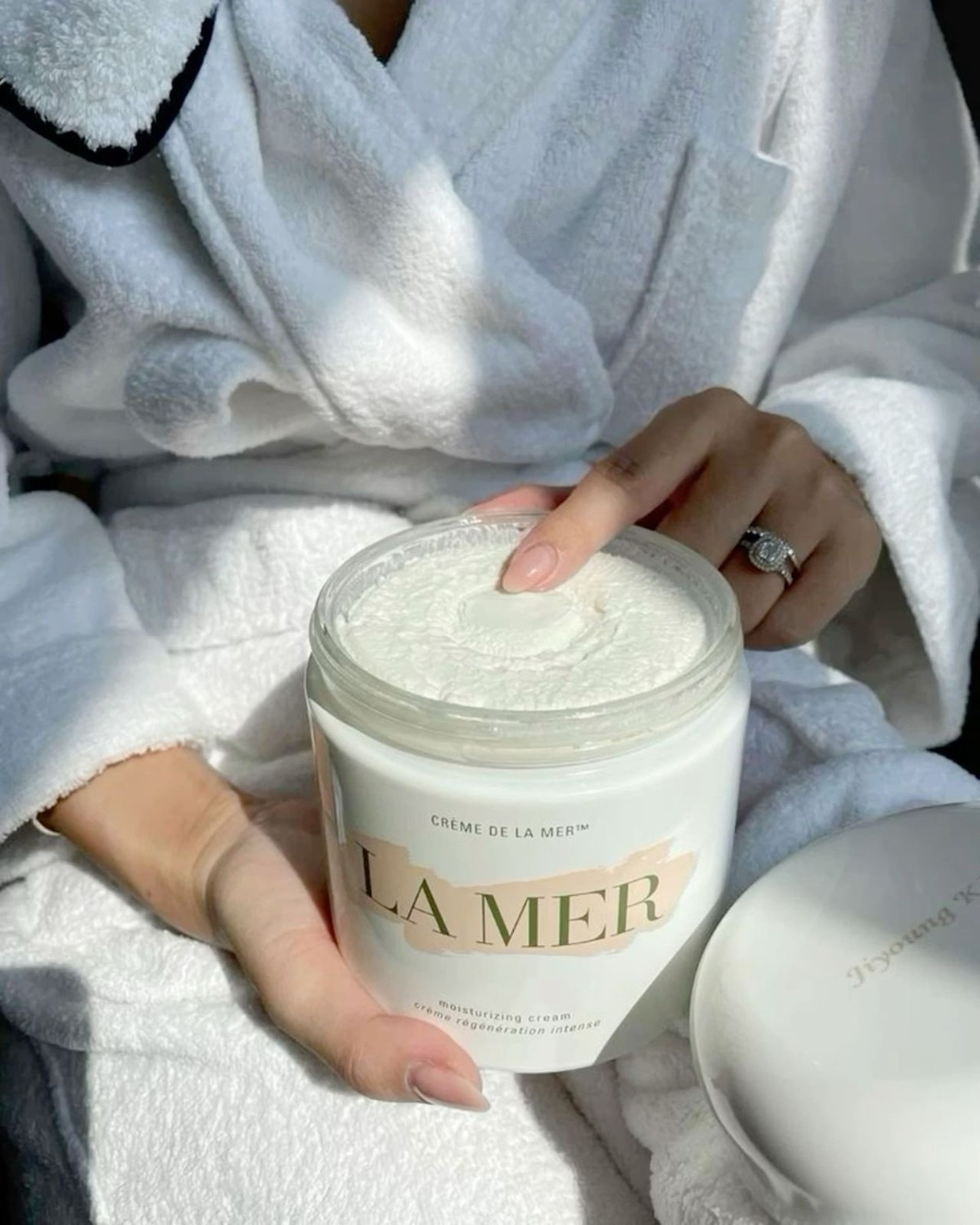 Woman in a white terry robe pressing a fingertip into a large open jar of Crème de la Mer moisturizing cream
