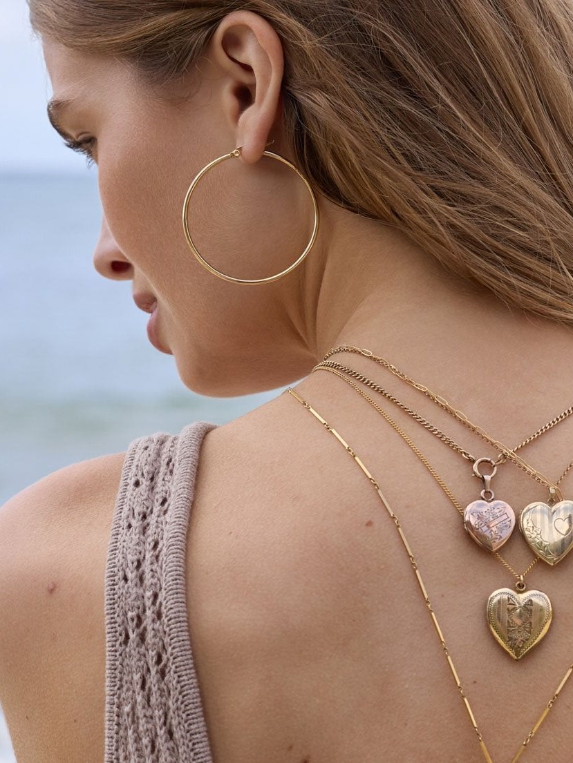 Woman in profile wearing a large hoop earring and layered heart necklaces against a pale beach backdrop