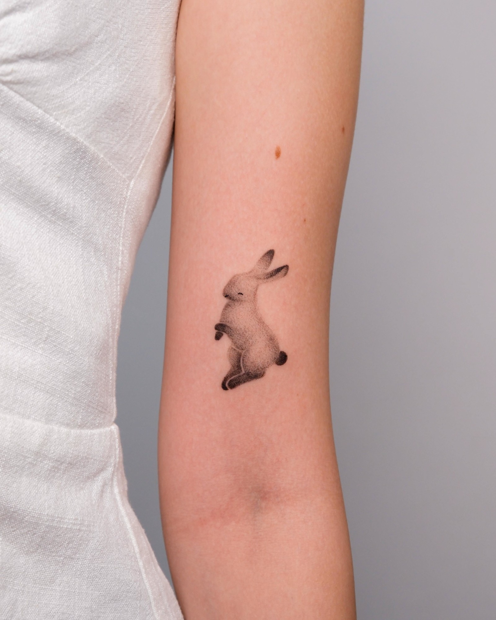 Small dotwork tattoo of a sitting rabbit on the back of an upper arm, next to a white linen top