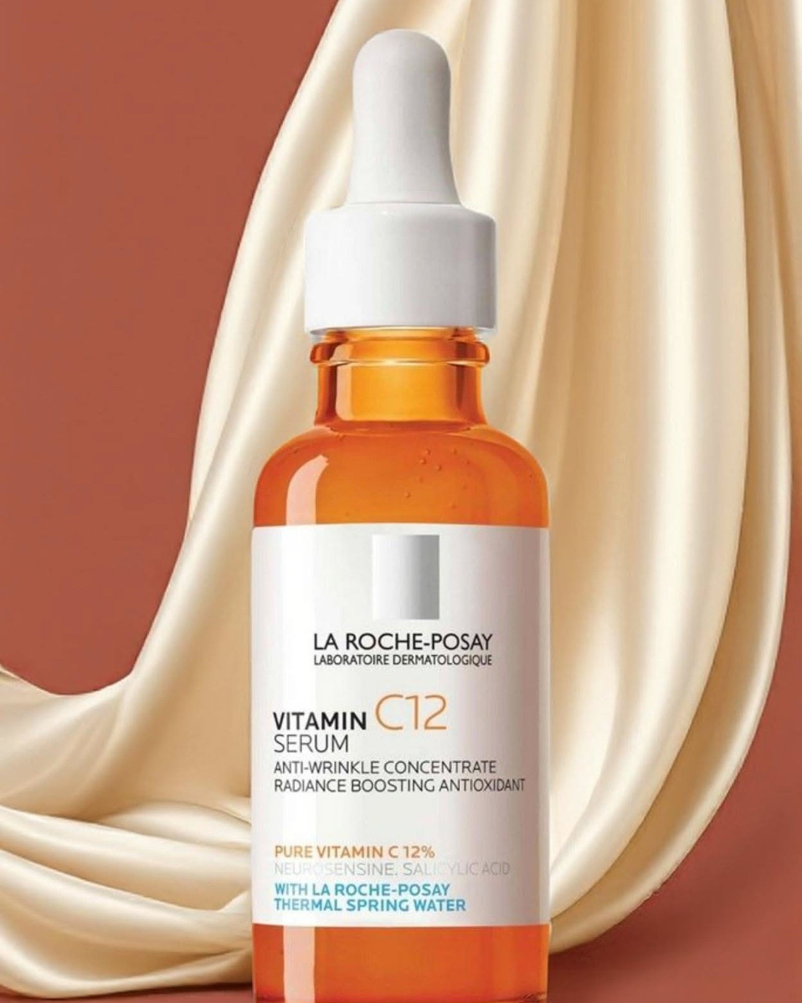 La Roche-Posay Vitamin C12 Serum in an orange glass dropper bottle in front of cream satin fabric on a terracotta background