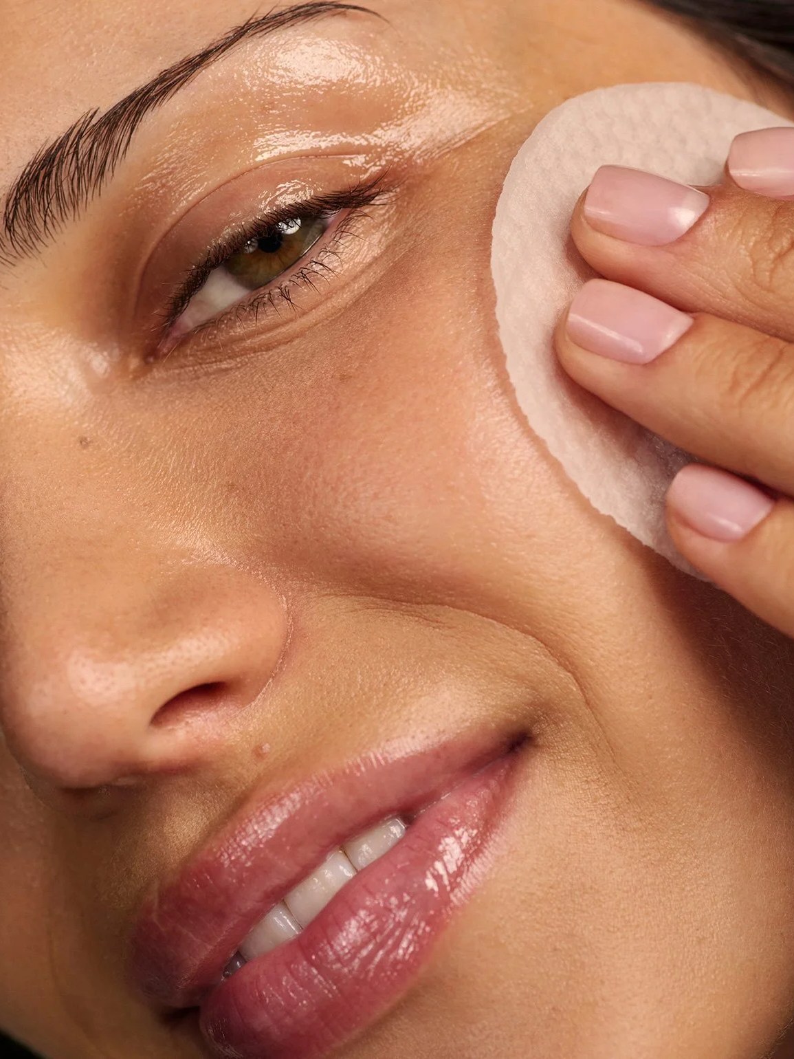 Model smiling while applying a cotton round near her eye in a close-up skincare campaign photo