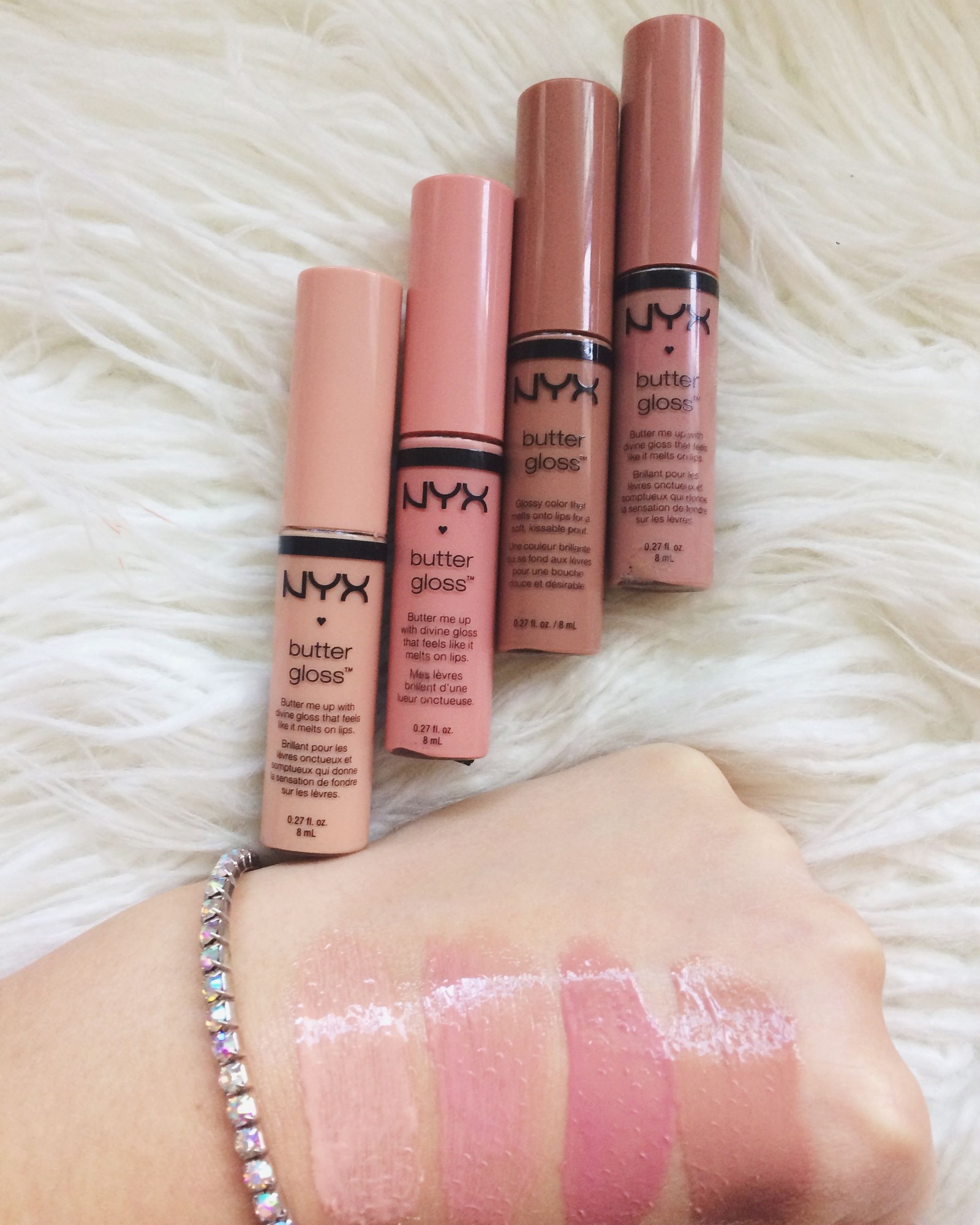Four NYX Butter Gloss tubes on a white faux fur rug above swatches on the back of a hand