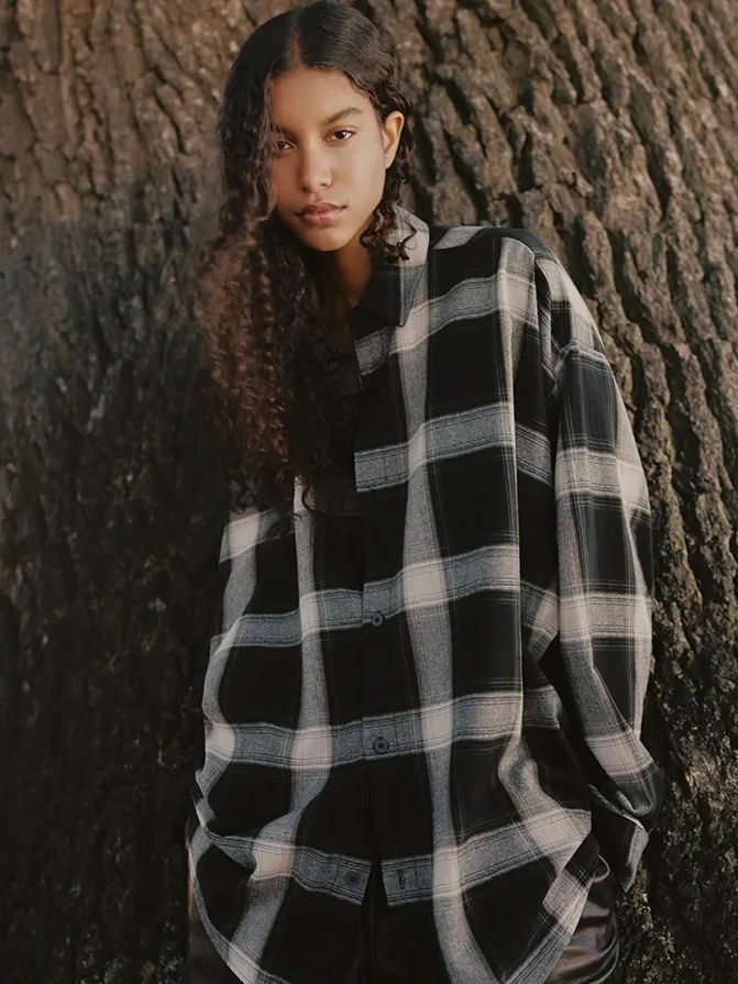 Woman in a black and white plaid shirt beside a tree in the ESSENTIALS Summer 2026 campaign