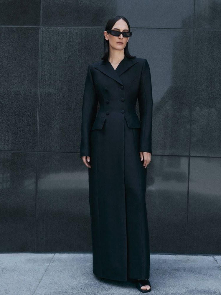Model in a long black tailored coat and sunglasses against dark stone panels