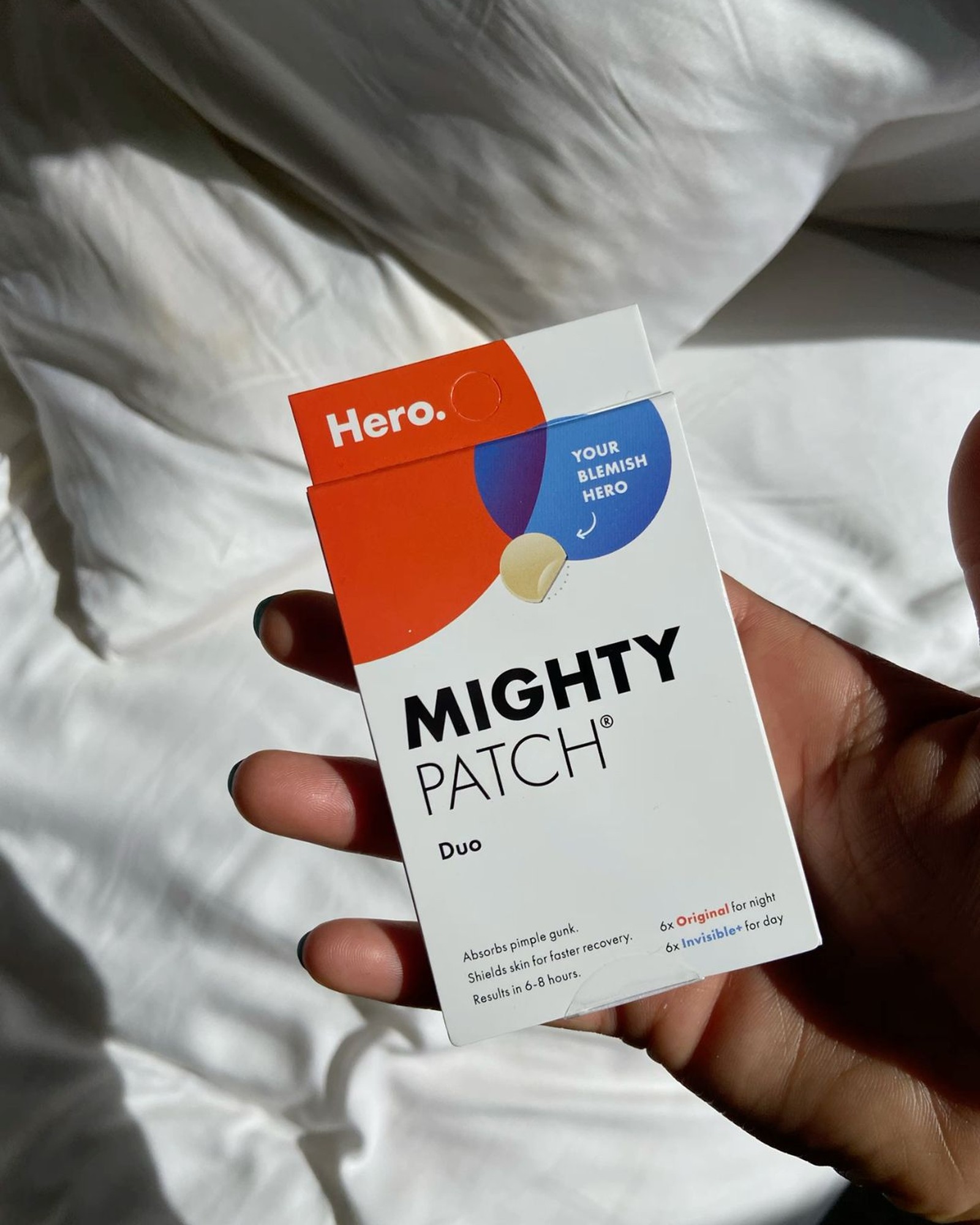 A hand holding a Hero Mighty Patch Duo box over white bed linen in sunlight