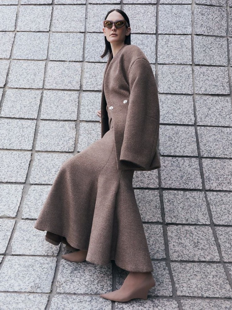 Model in a sculptural taupe coat and amber sunglasses beside a patterned stone wall
