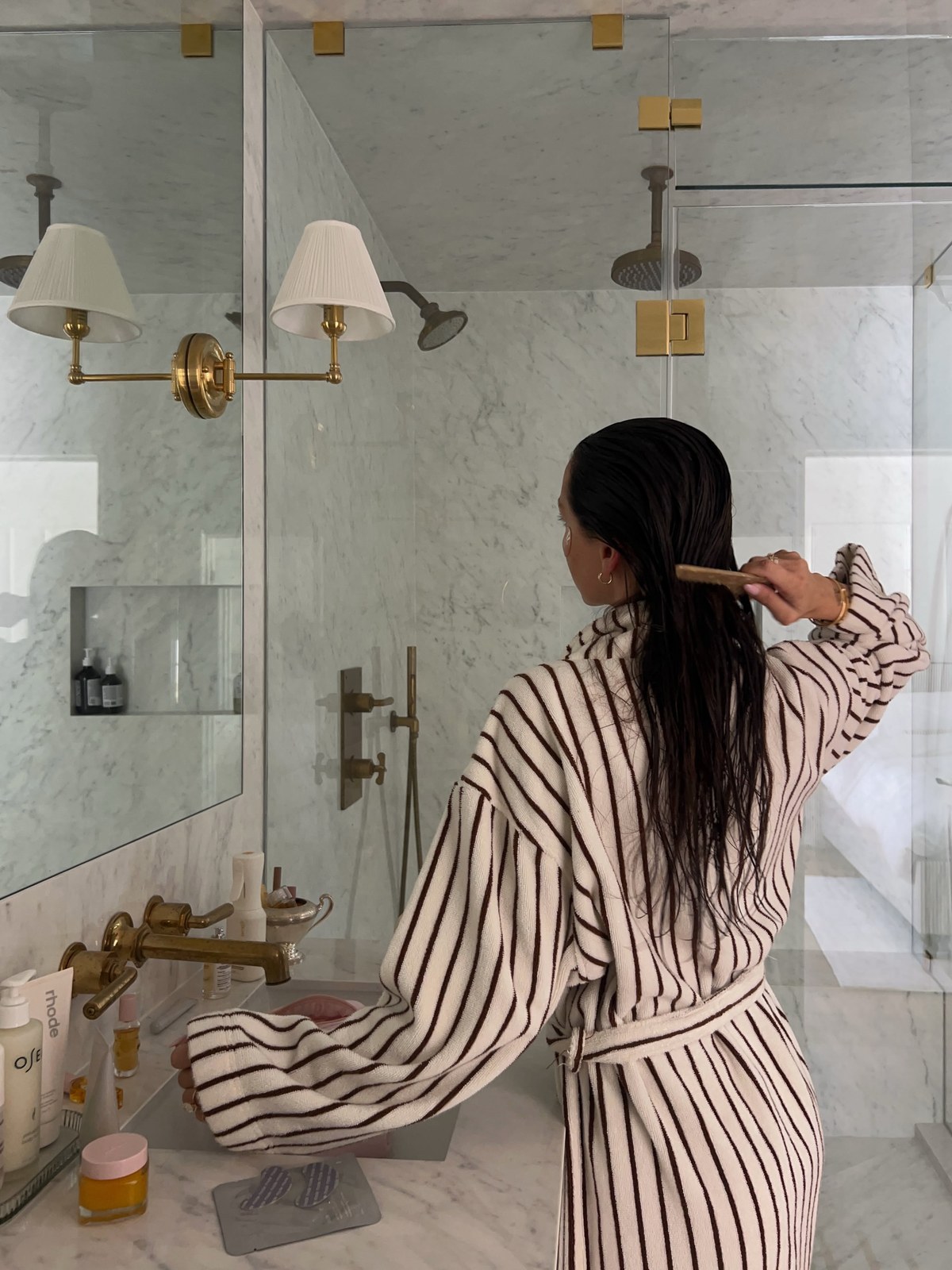 Woman from behind in a striped robe combing wet hair at a marble bathroom vanity with brass fittings