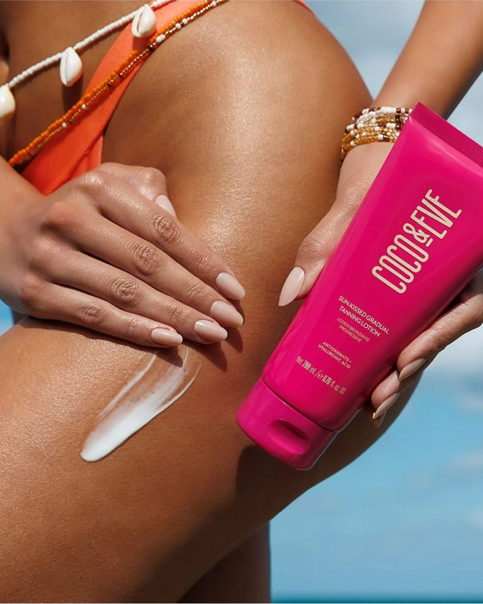 Tanned leg with a swipe of white lotion, a hand holding a pink Coco & Eve Sun-Kissed Gradual Tanning Lotion tube, and a turquoise sea behind