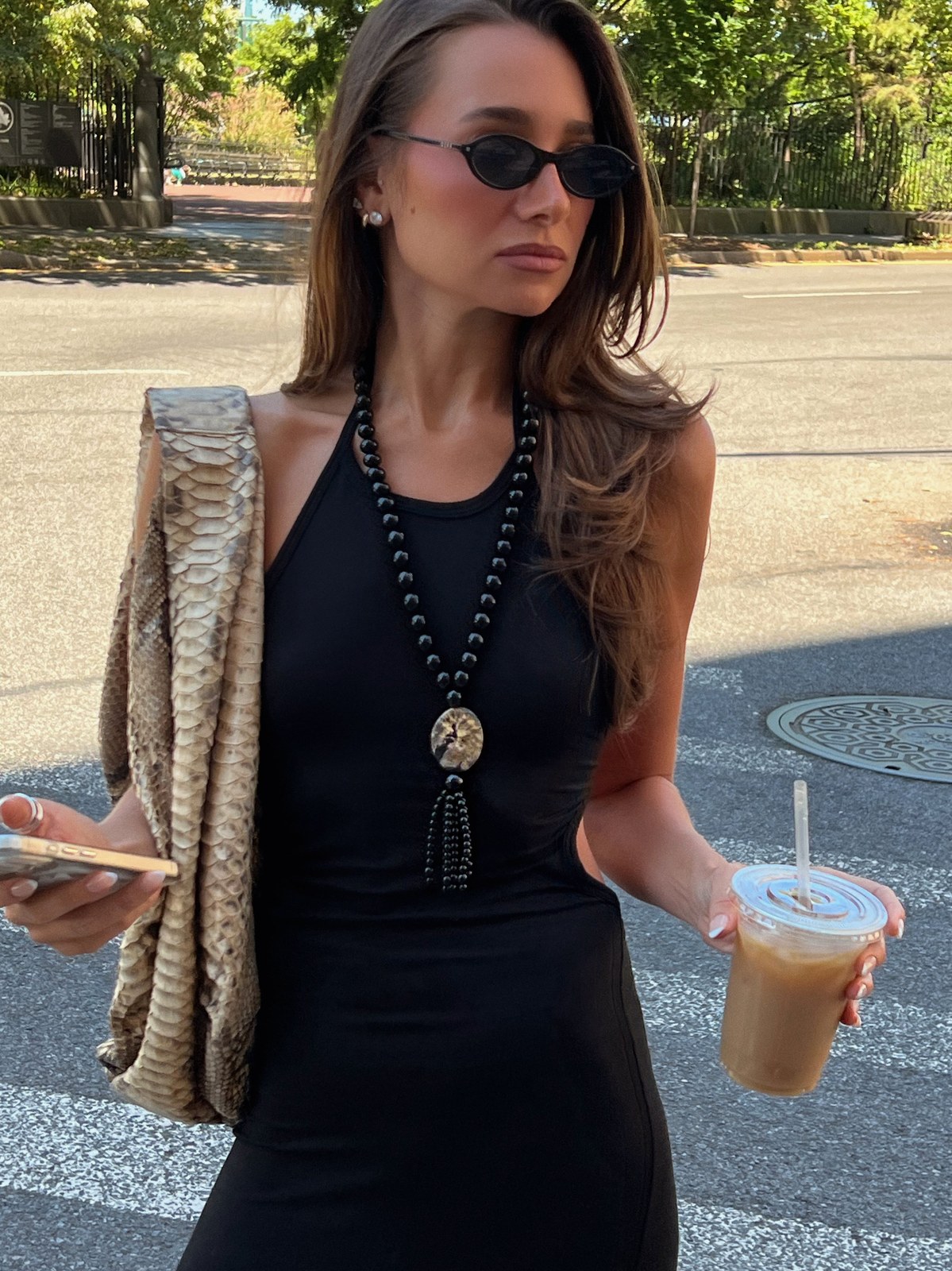 Hannah B in a black fitted dress with sunglasses on a tree-lined sidewalk holding a drink and knitwear