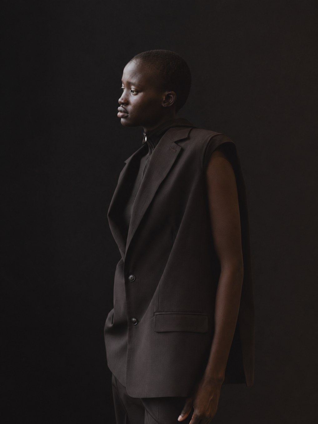 Model in a dark brown sleeveless tailored suit against a black studio backdrop