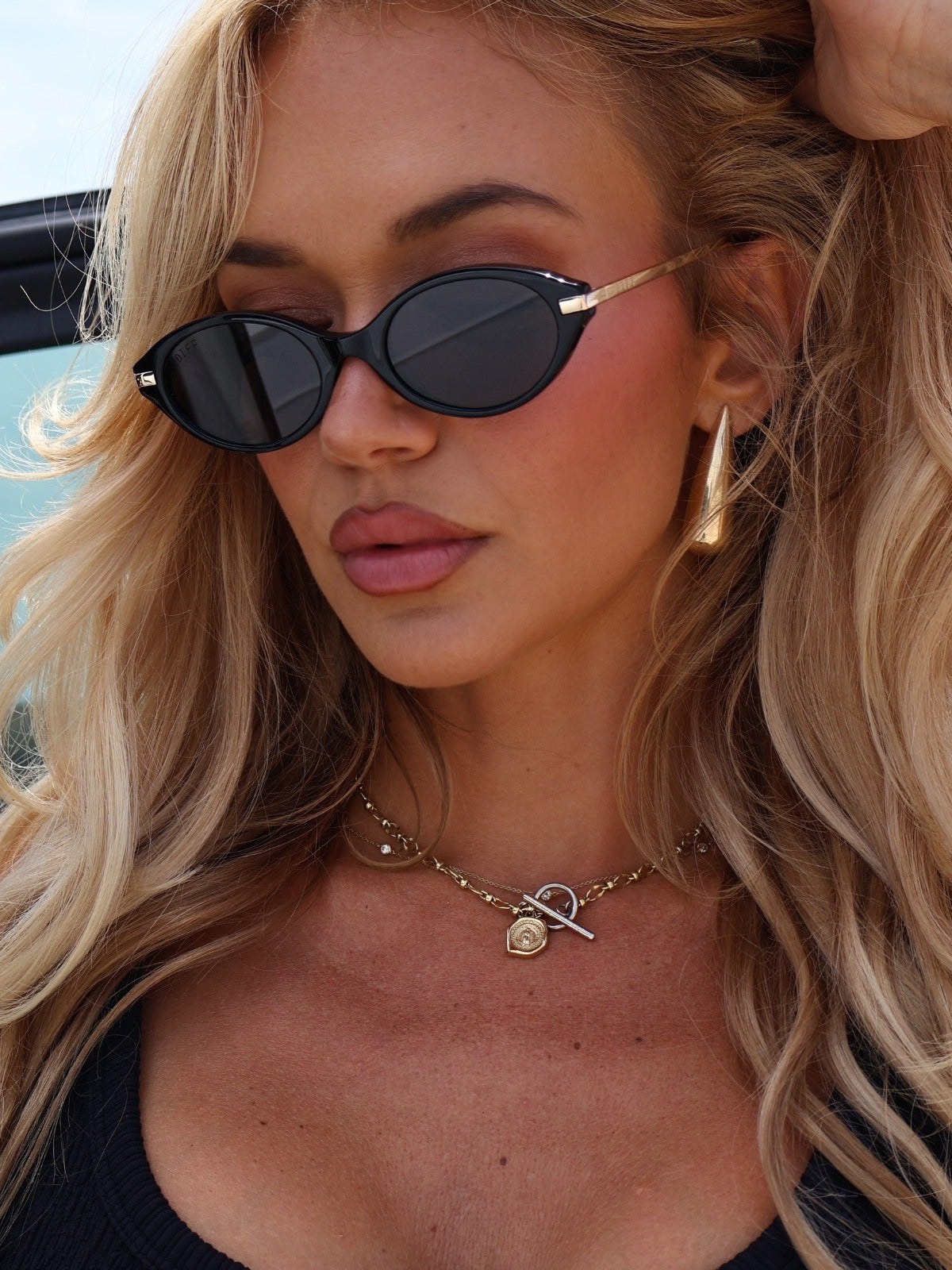 Blonde model wearing black oval DIFF sunglasses beside a bright blue backdrop