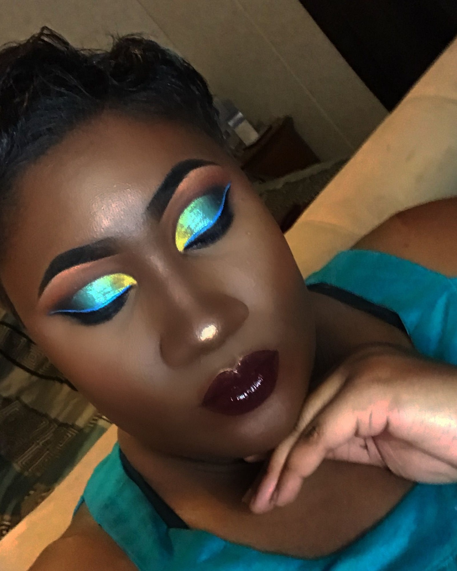 Woman with short dark hair in a teal top, wearing teal and gold chrome eyeshadow with bright blue liner and a deep plum glossy lip