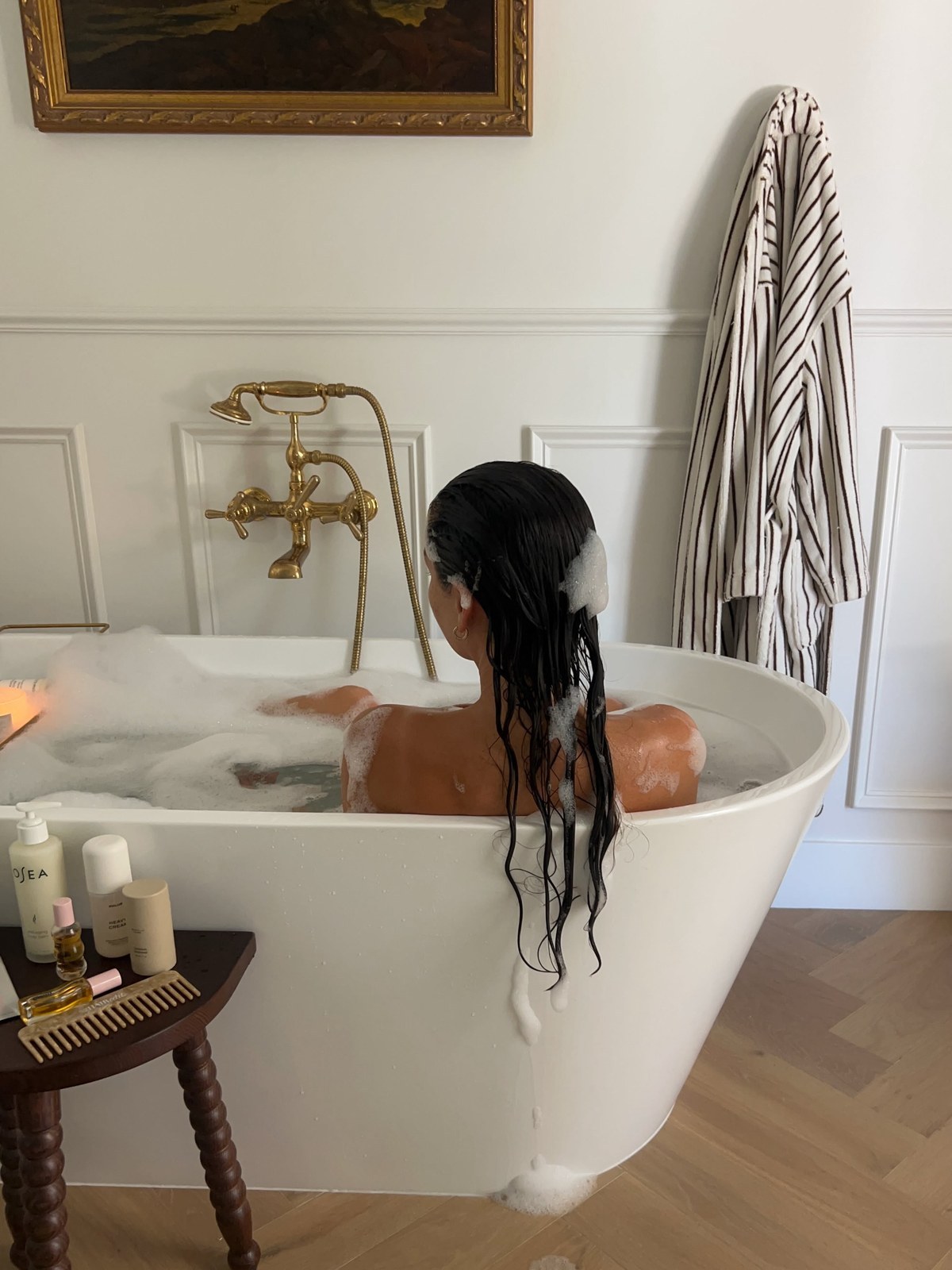 Woman from behind in a bubble bath with a striped robe on the wall and a small table of body products beside the tub