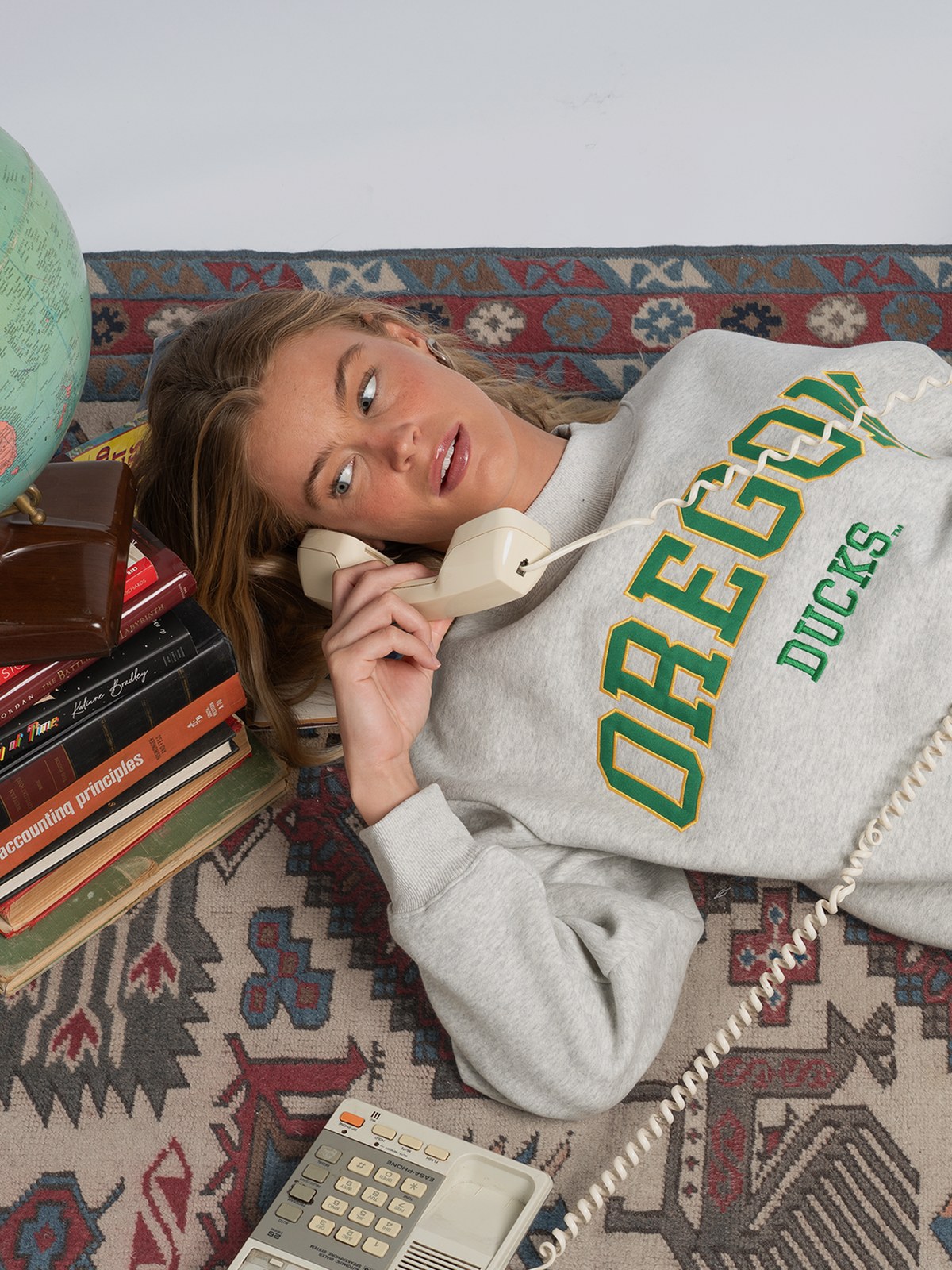 Model in an Oregon Ducks sweatshirt reclining beside books and a globe