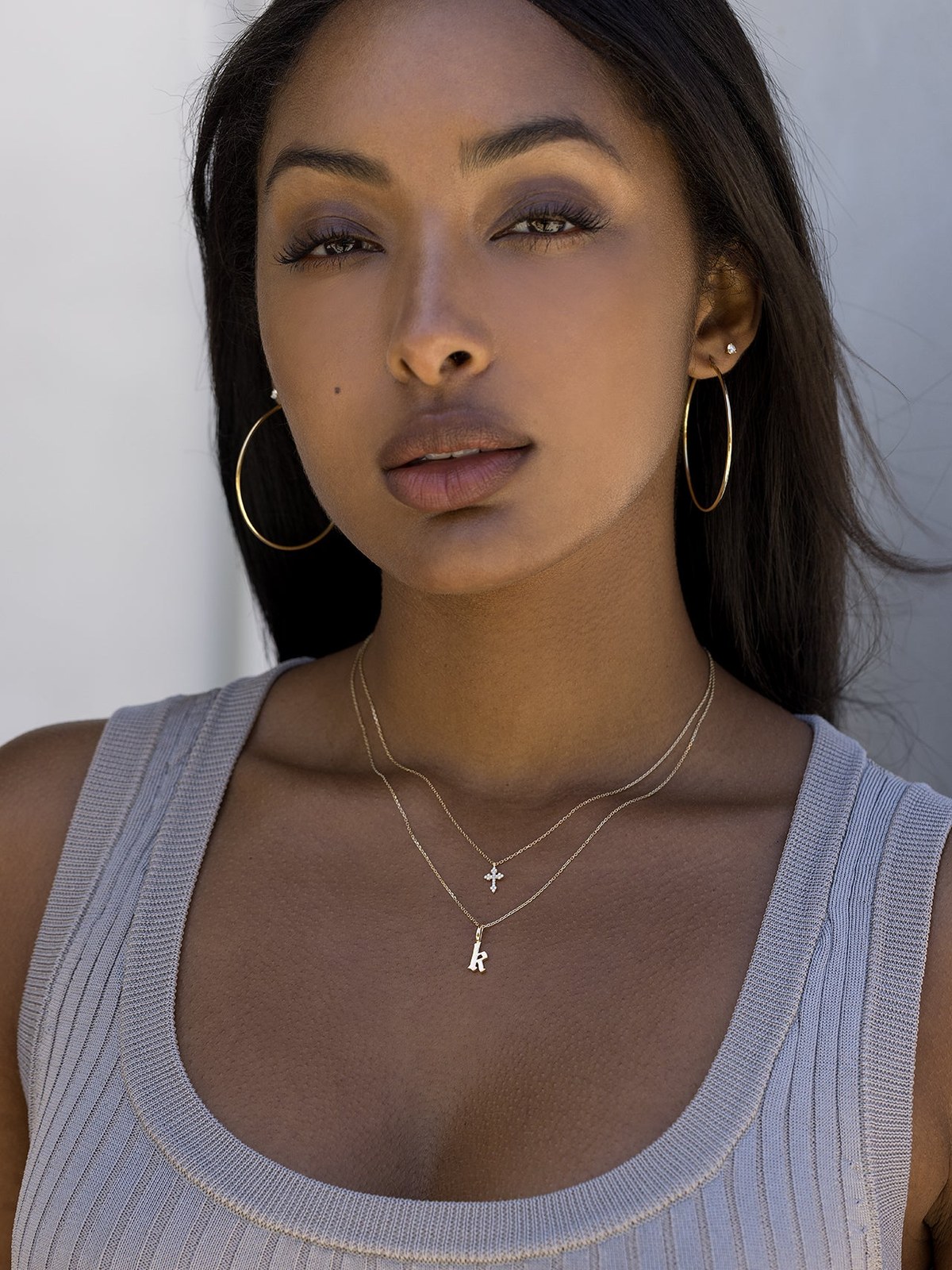 Model with long dark hair wearing gold hoops and a layered necklace against a soft gray backdrop