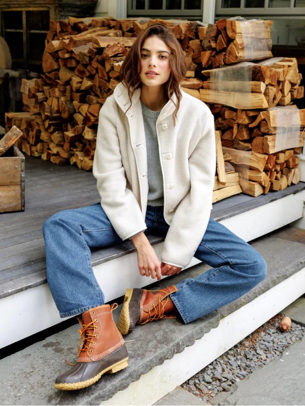 Woman in a pale sherpa layer and jeans seated beside stacked firewood