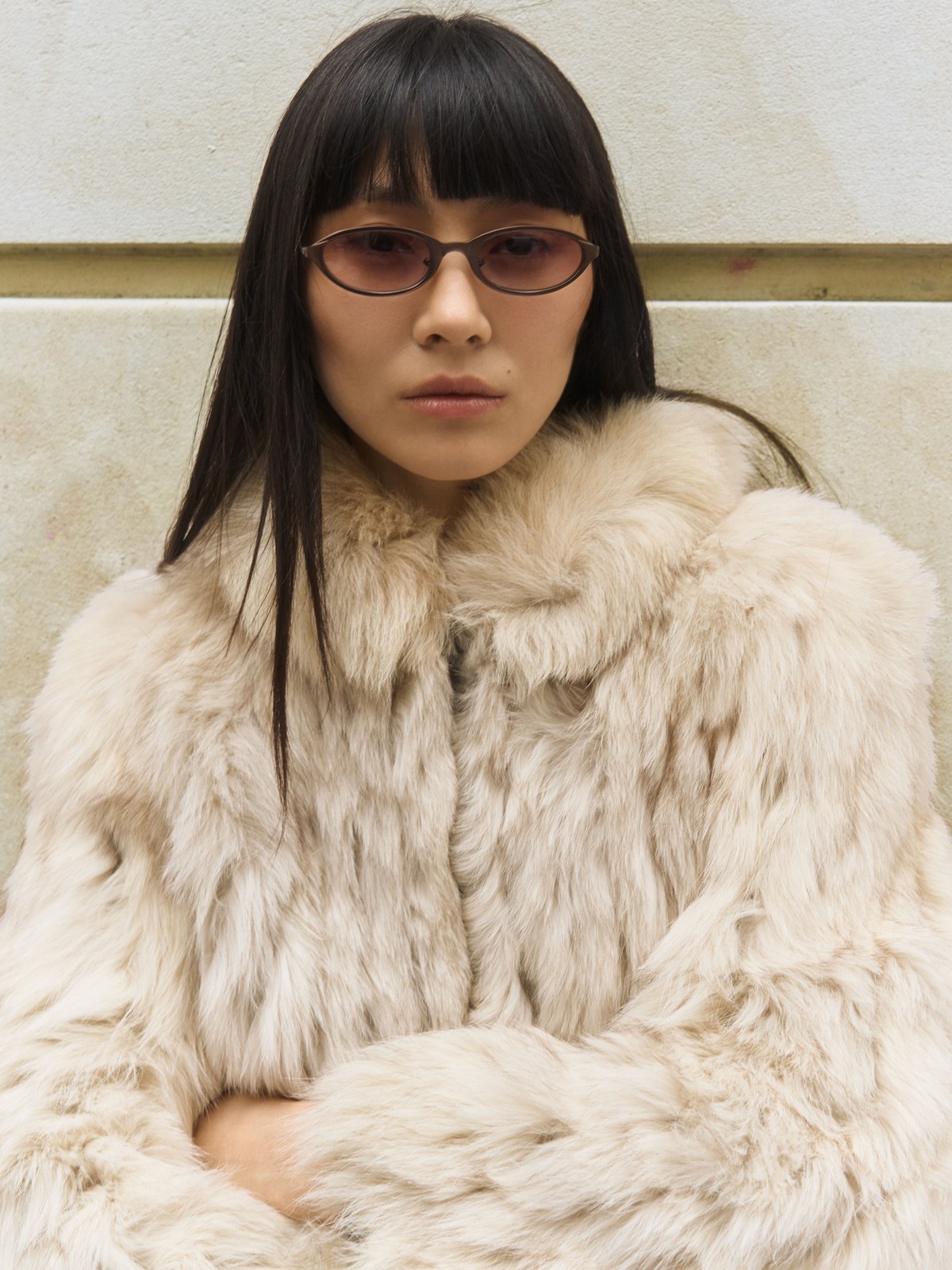Woman in BRU sunglasses and a pale textured coat against a stone wall