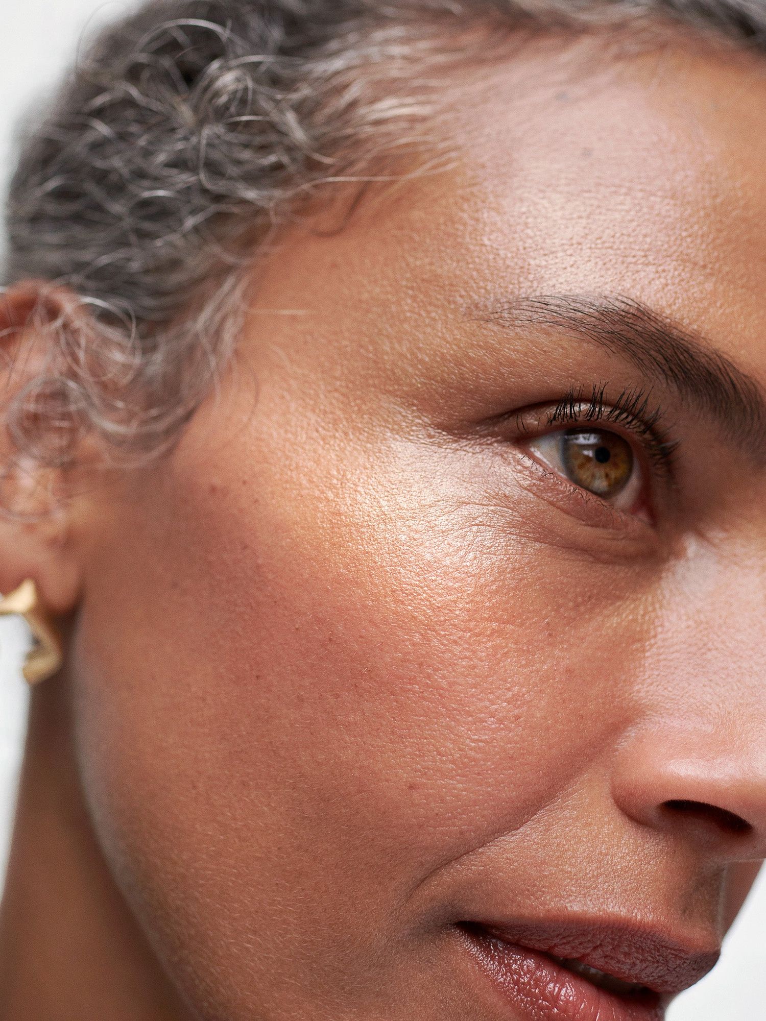 Woman with grey hair and a glowing highlighted cheekbone wearing a gold earring