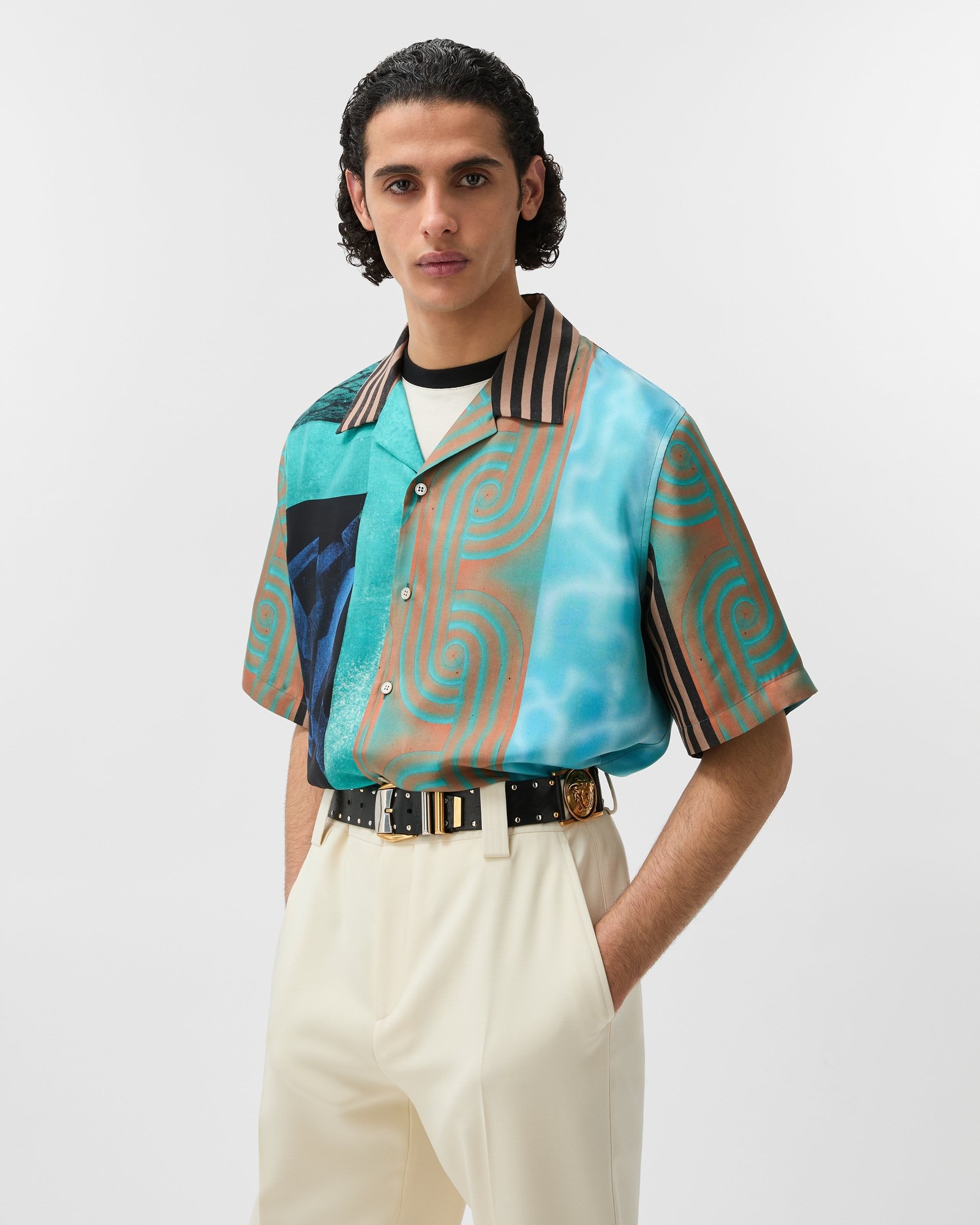 Man with dark curly hair in a turquoise and copper printed camp-collar shirt, black studded belt and cream trousers