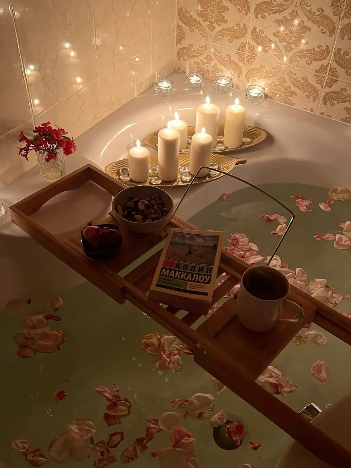 Candlelit bathtub with petals floating in the water, a wooden bath tray holding a book, a mug and a bowl of nuts, and pillar candles on a gold leaf-shaped dish
