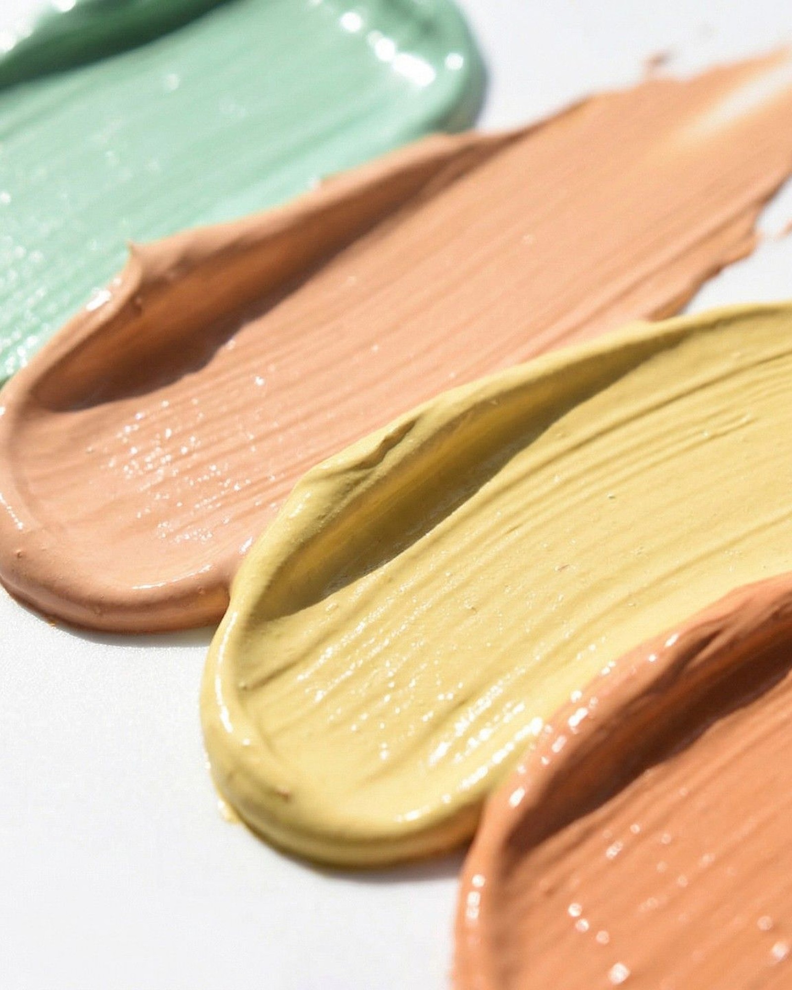 Close-up swatches of green, peach and yellow color corrector creams on a white surface