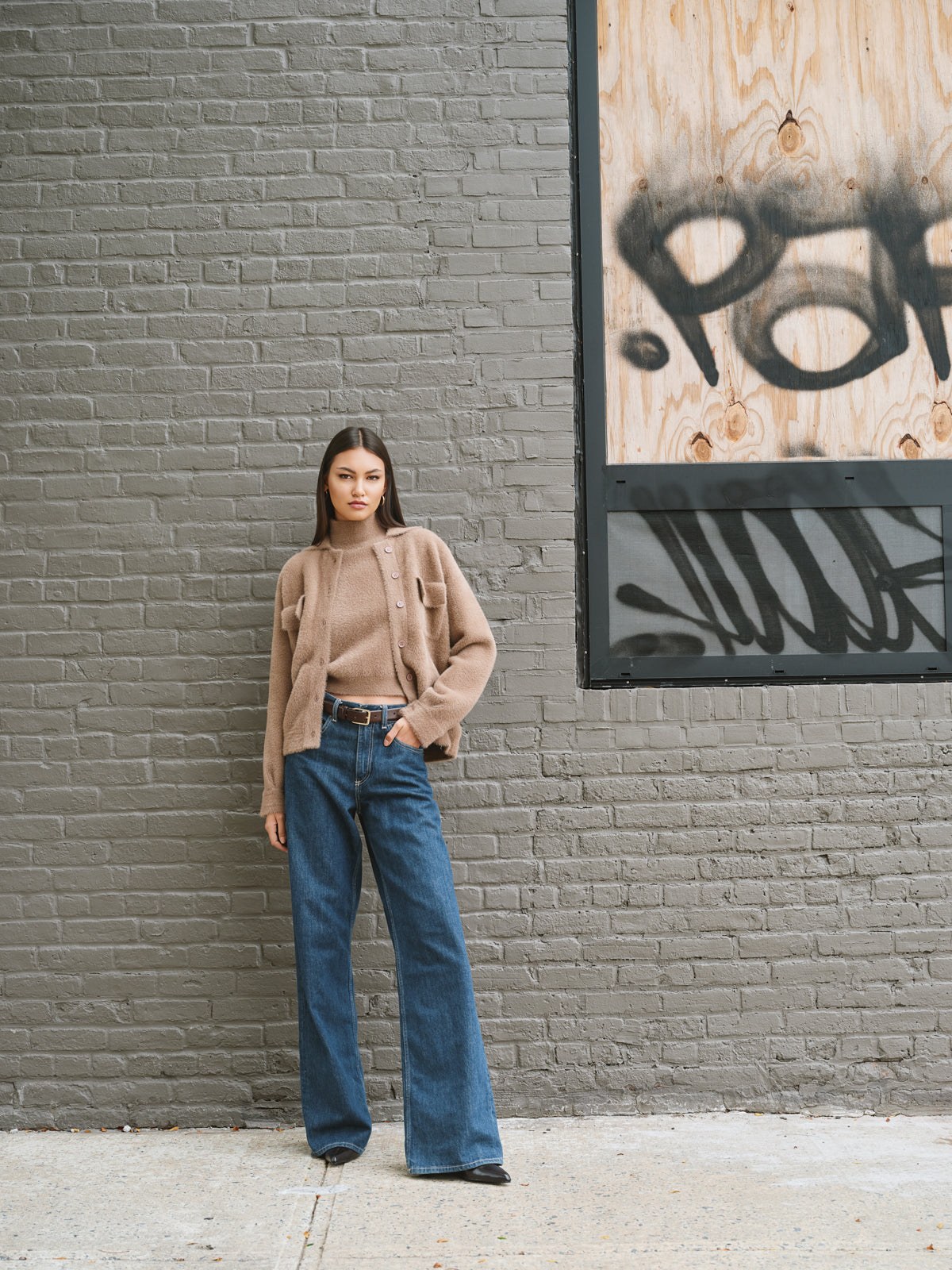 Model in a warm neutral knit and jeans against a graffiti-marked brick wall