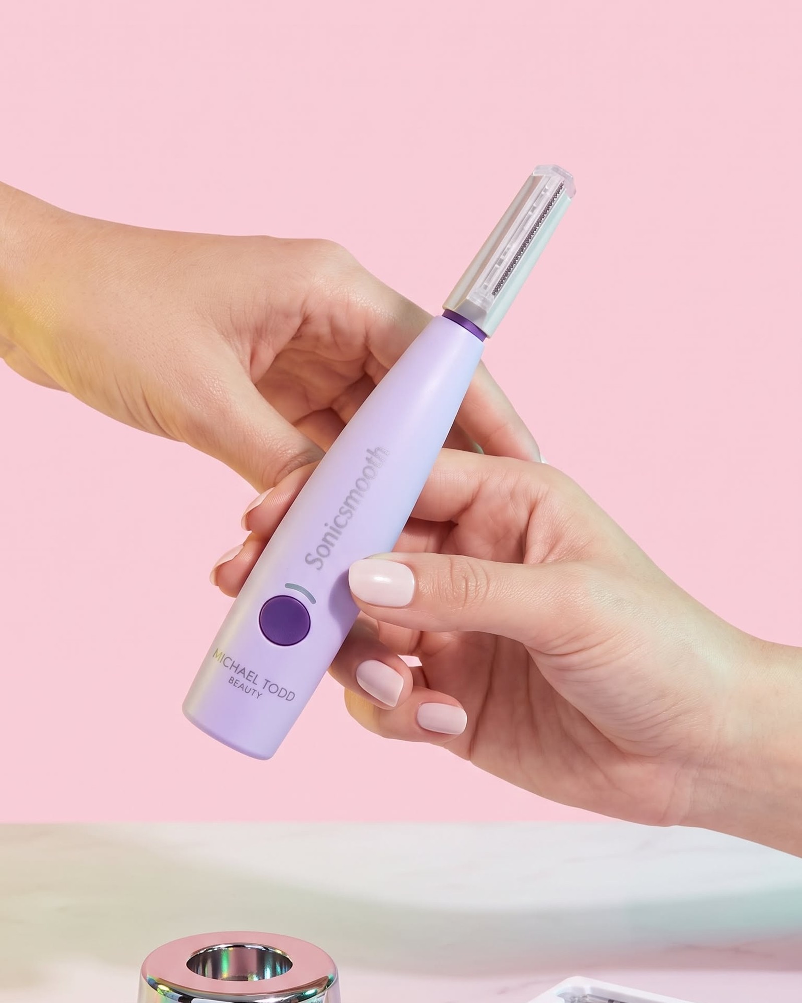 Two hands passing a lilac Michael Todd Beauty Sonicsmooth dermaplaning wand with a clear blade cover against a soft pink background