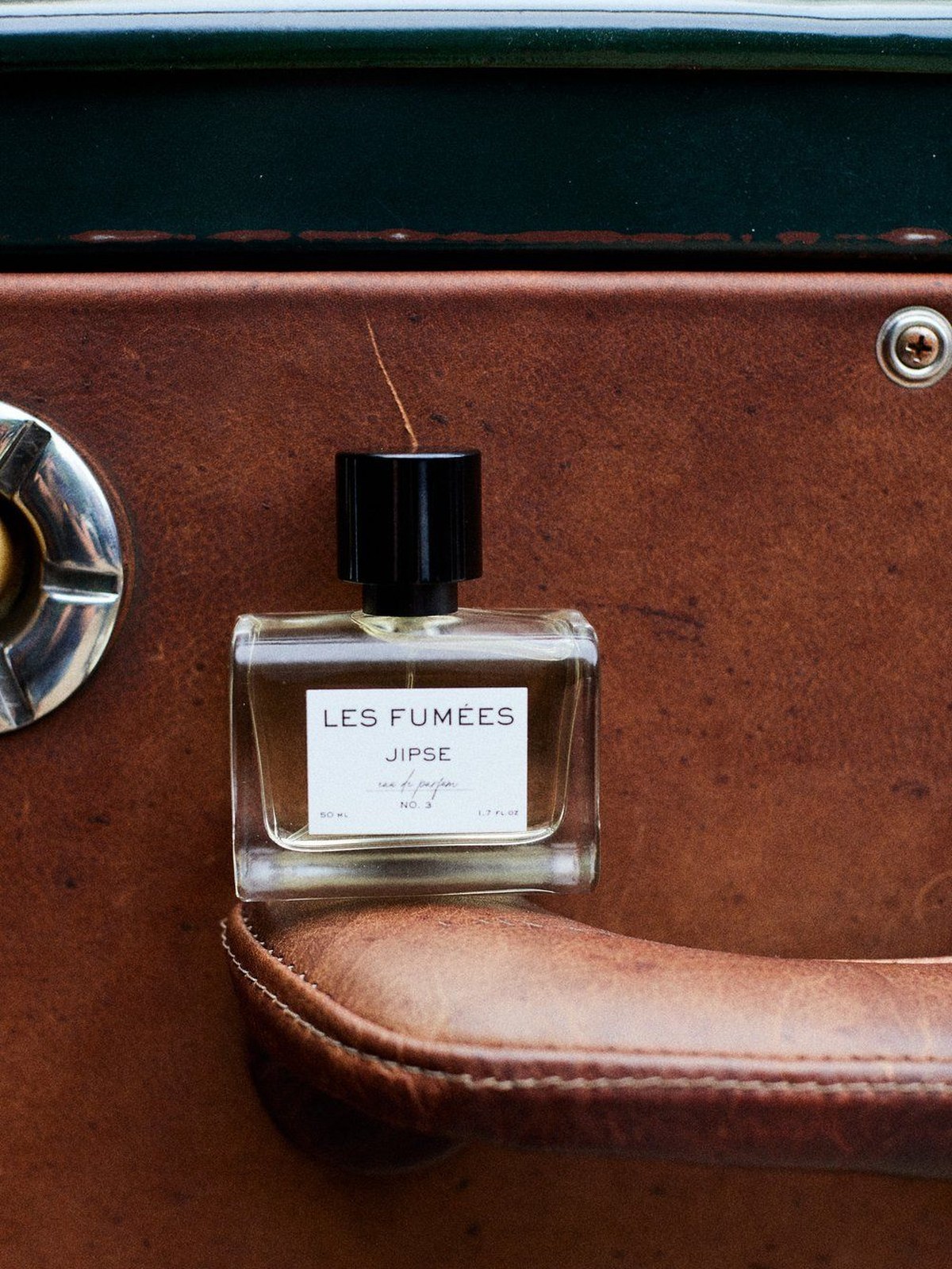 Bottle of Les Fumées Jipse eau de parfum resting on a tan leather car door handle