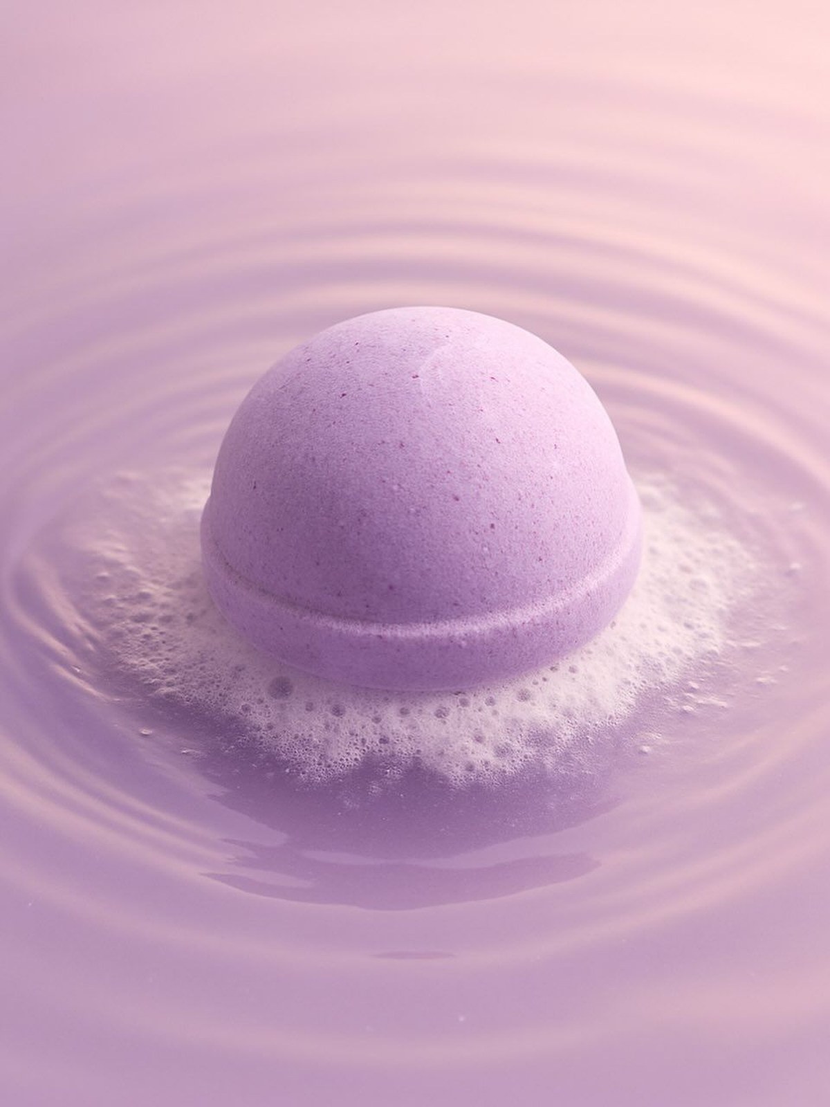 A lilac bath bomb fizzing in lilac water, with rings rippling out from it and a soft white foam around its edge.