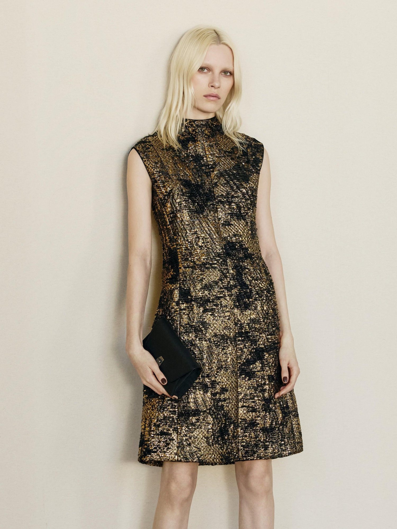 Model in a sleeveless black and gold jacquard dress holding a clutch against a beige wall