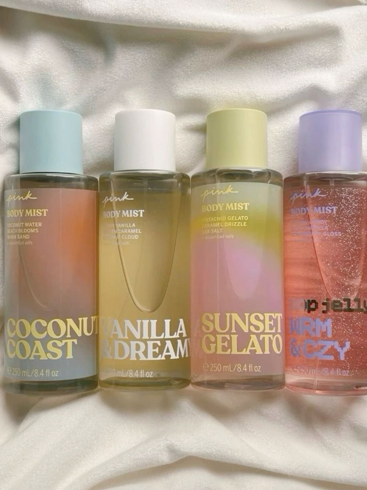 Four pastel body mist bottles in Coconut Coast, Vanilla & Dream, Sunset Gelato and a glittery pink shade lined up on white fabric