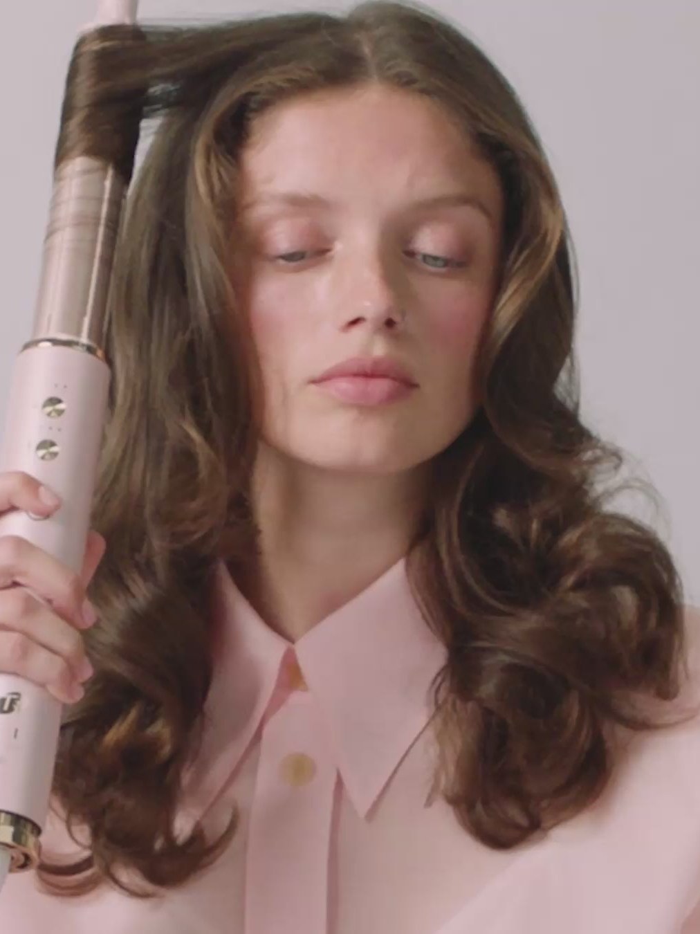 Model with glossy brown curls holding a white curling tool in a pale studio