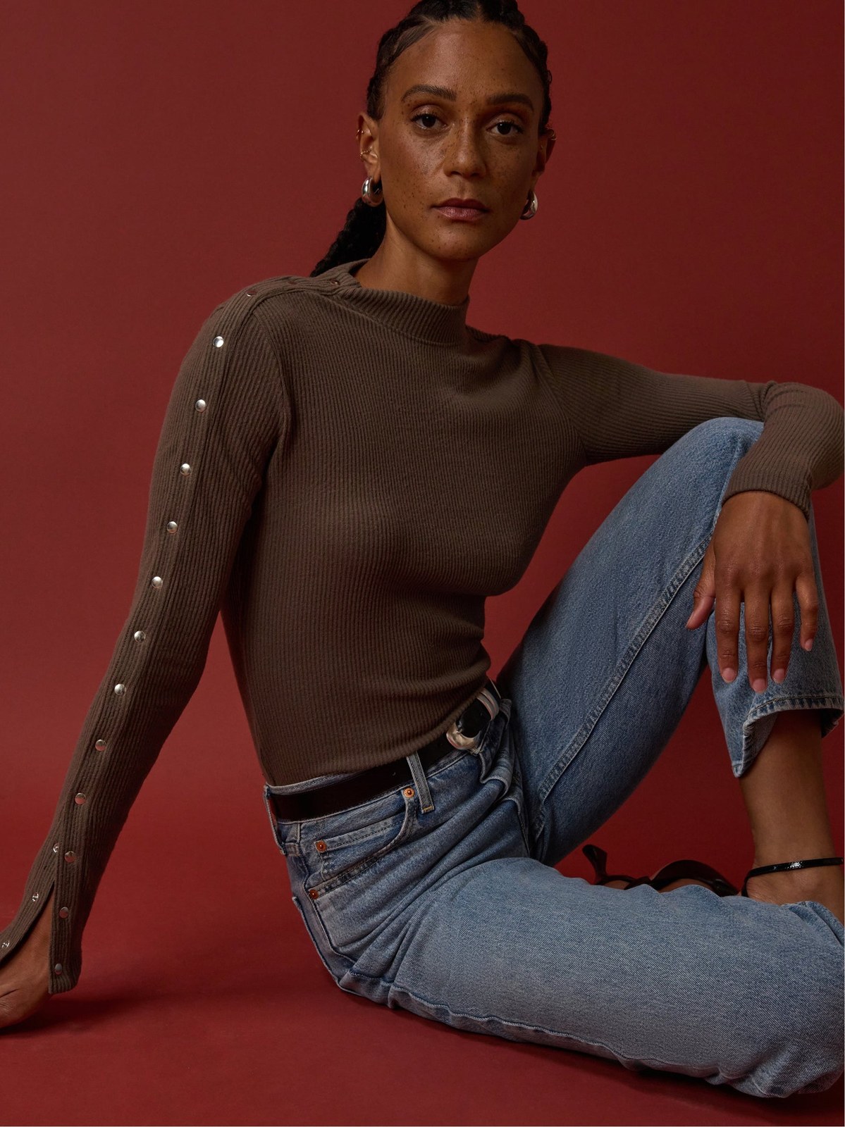 Model seated in a brown knit top and jeans against a red studio backdrop