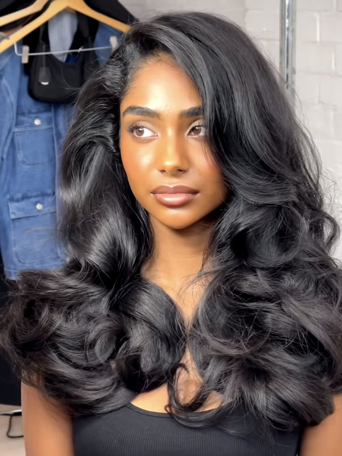 Woman with long glossy black voluminous waves and a side part, standing in front of a brick wall with denim jackets on hangers