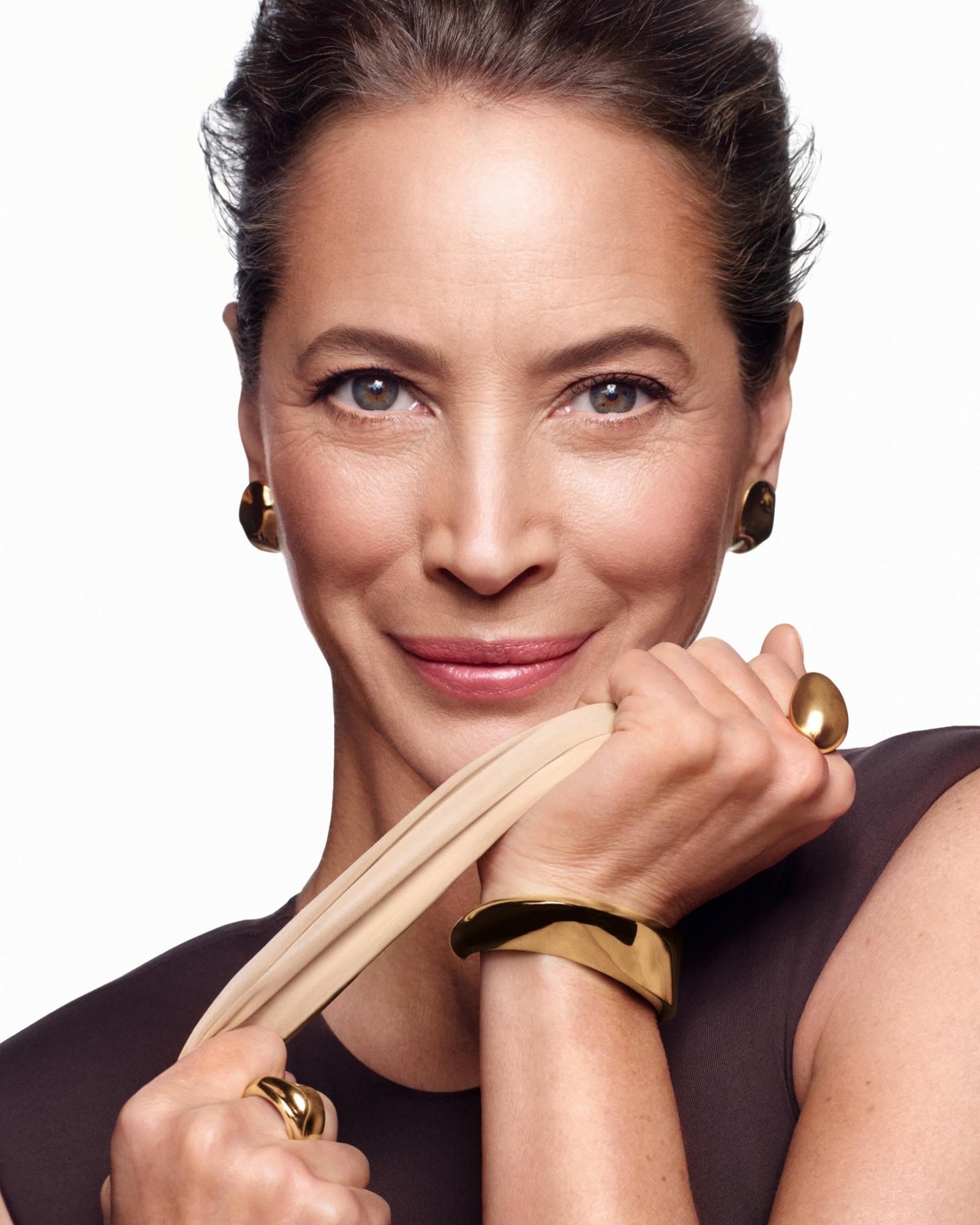 Smiling woman with a dark hair updo wearing gold sculptural earrings, cuff and rings, holding a beige stretch band against a white background