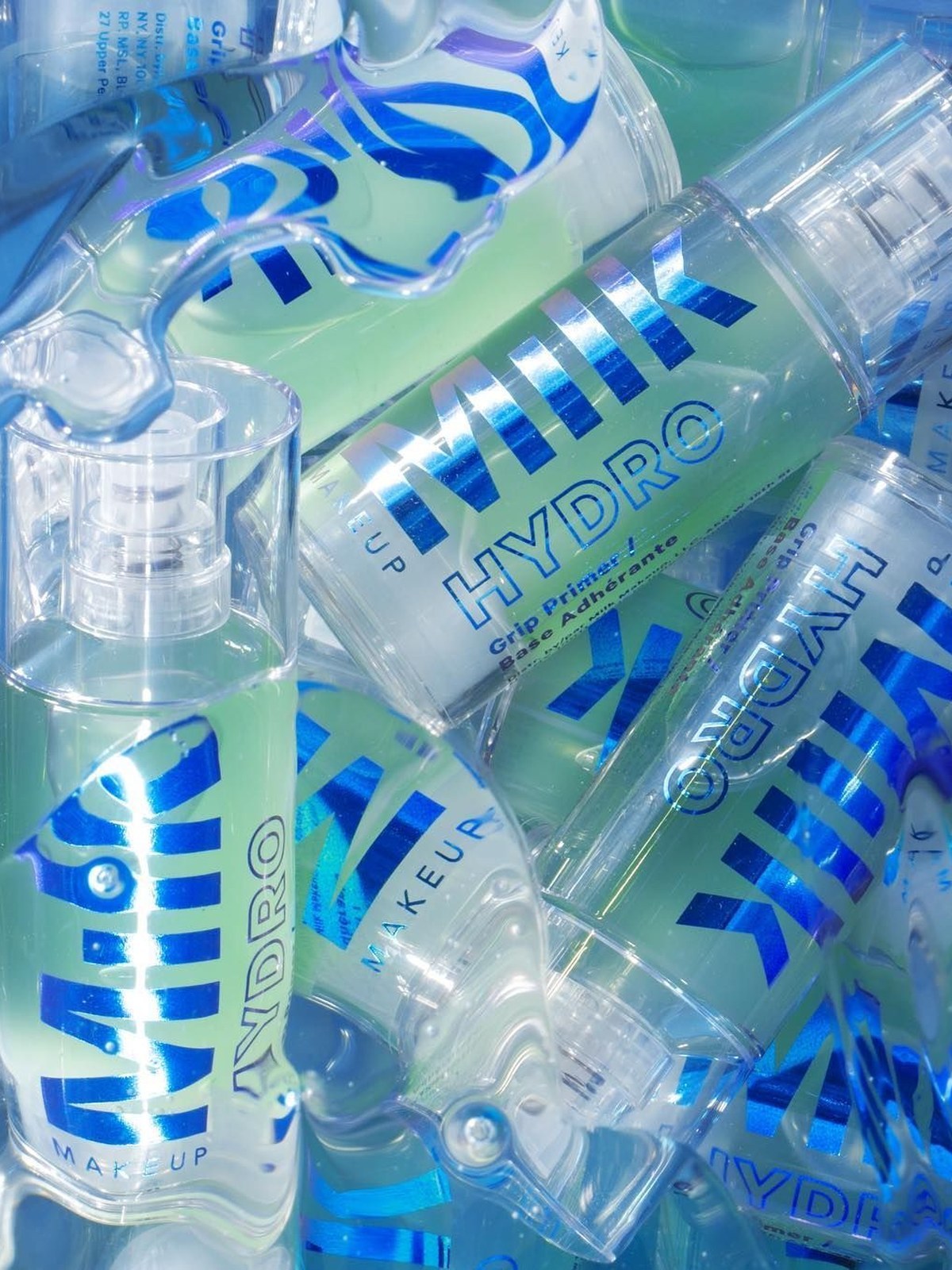 Pile of clear Milk Makeup Hydro Grip bottles and primer sticks with holographic blue logos, some covered in water