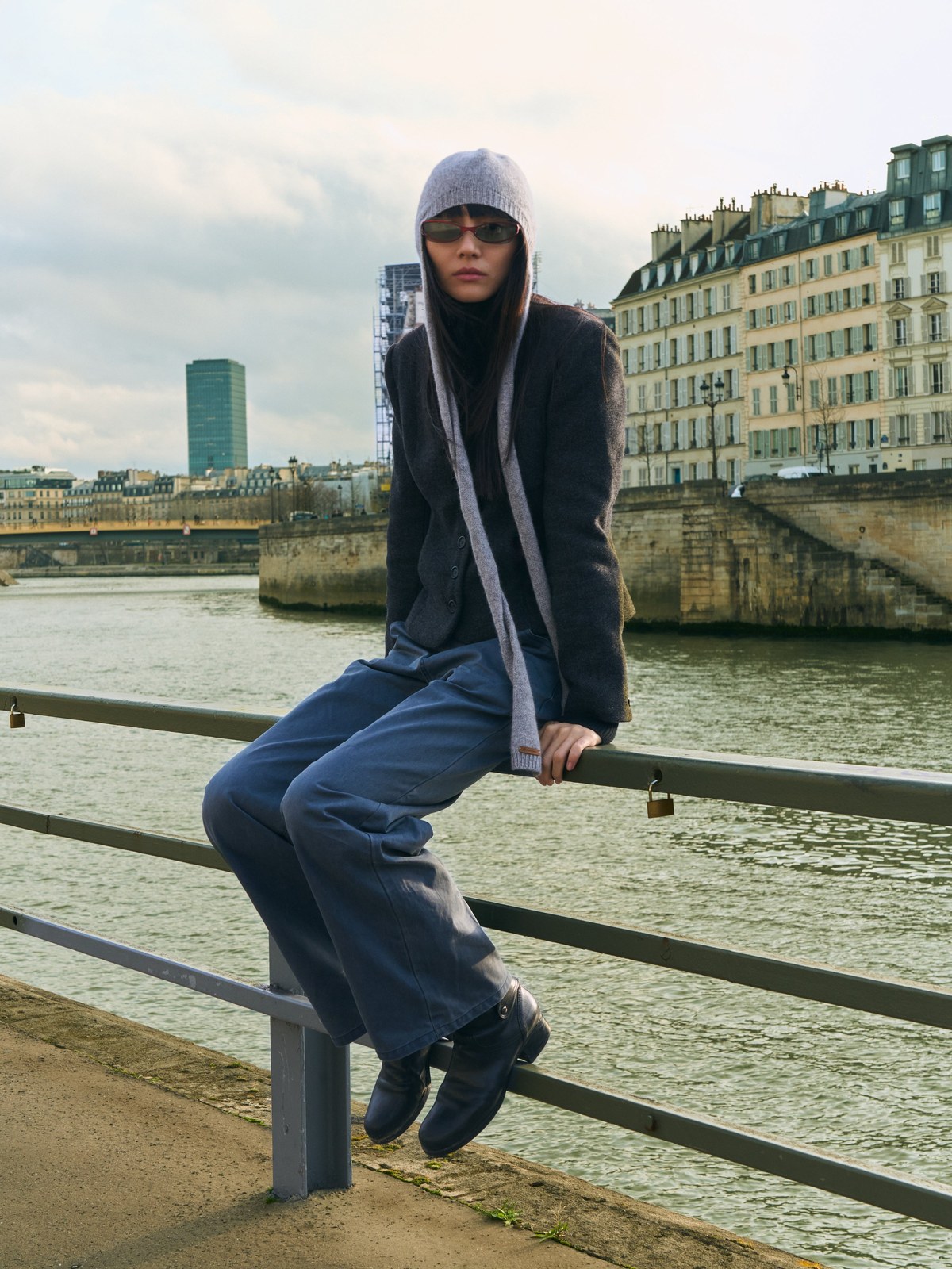 Model in BRU sunglasses seated on a riverside railing with Paris buildings behind her