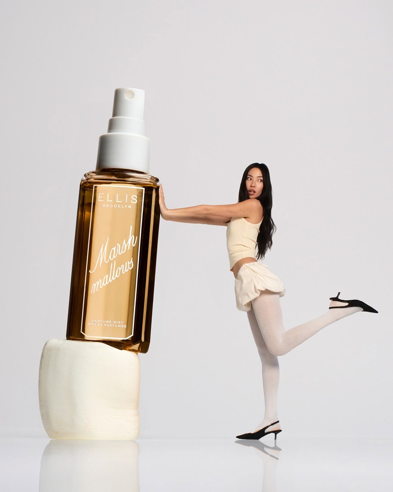 Woman in cream top, skirt and white tights leaning on a giant Ellis Brooklyn Marshmallows perfume mist bottle balanced on a marshmallow