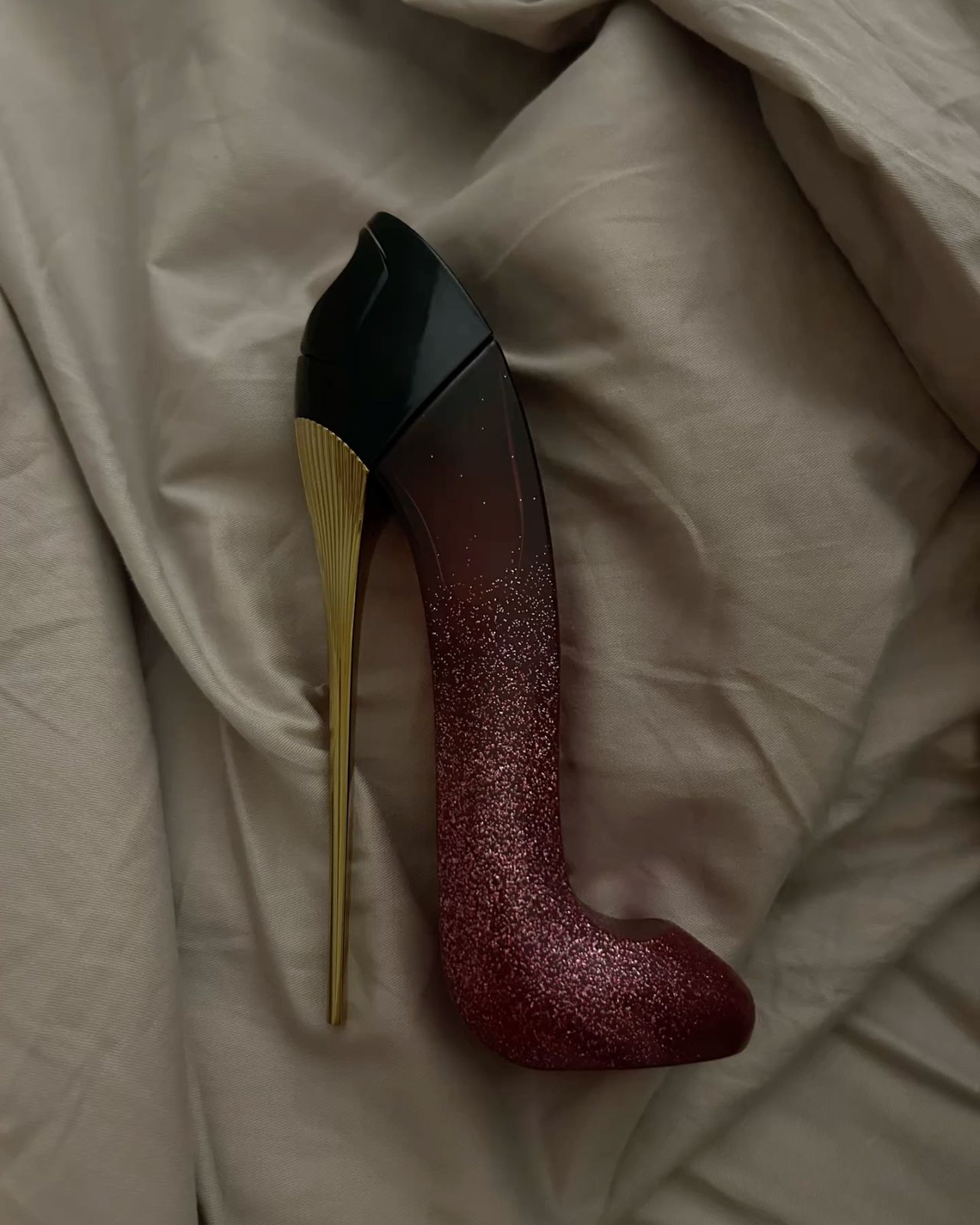 Stiletto-shaped perfume bottle with a glittery red to black gradient and a gold heel lying on rumpled beige fabric