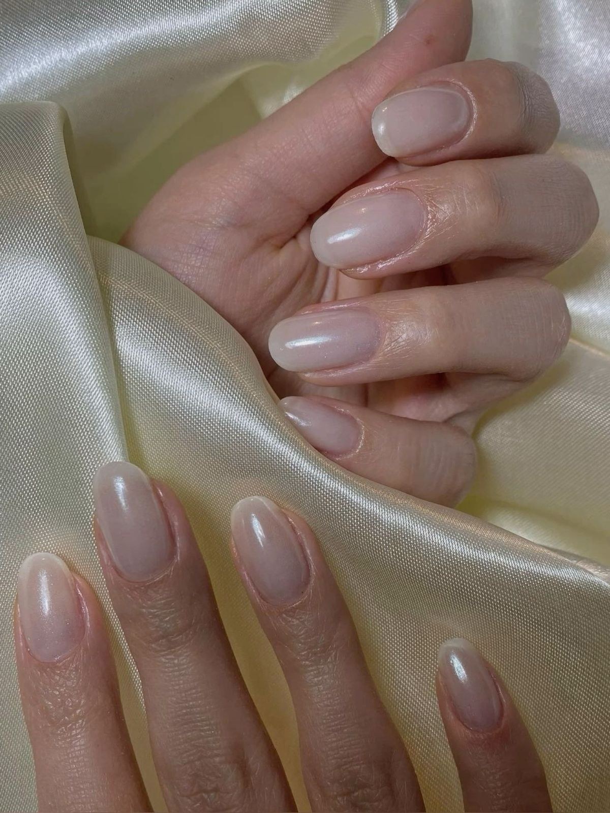 Two hands with oval nails in a pearly sheer nude polish resting on pale satin