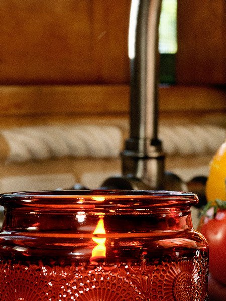 A lit red glass candle beside tomatoes and a hand at a kitchen counter