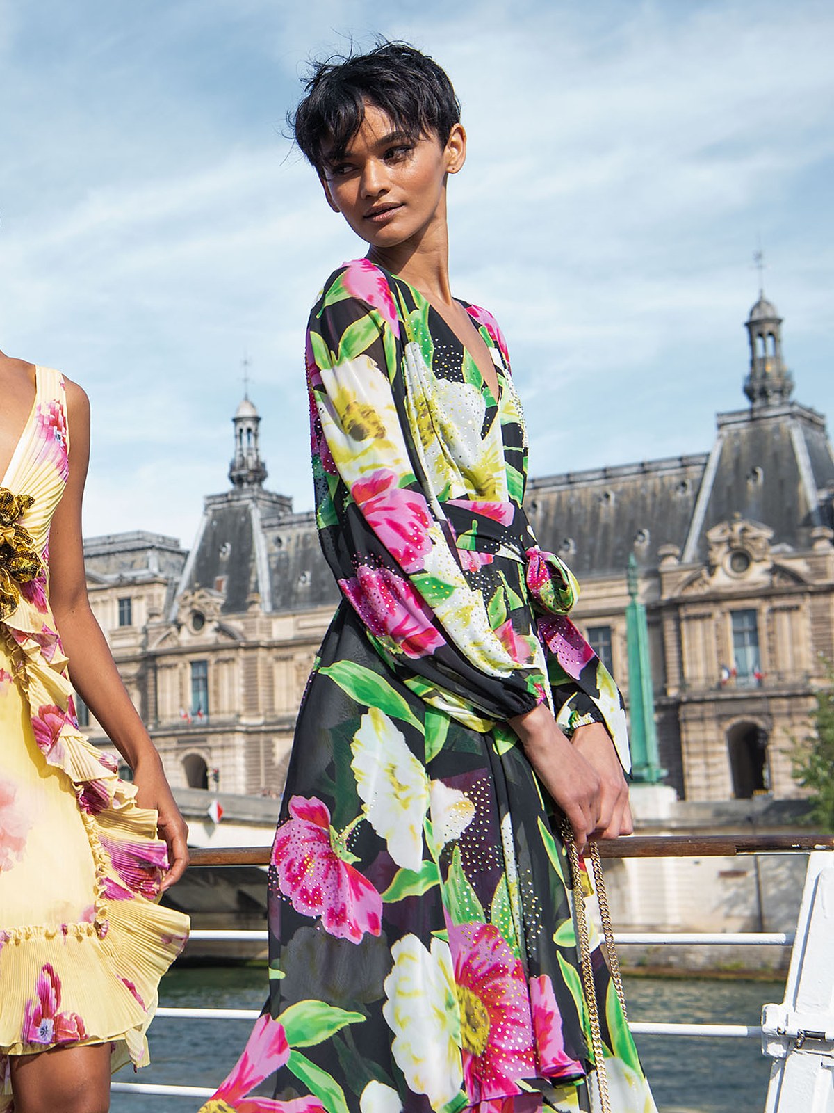 Models in bold floral print dresses on the Seine with Paris rooftops behind